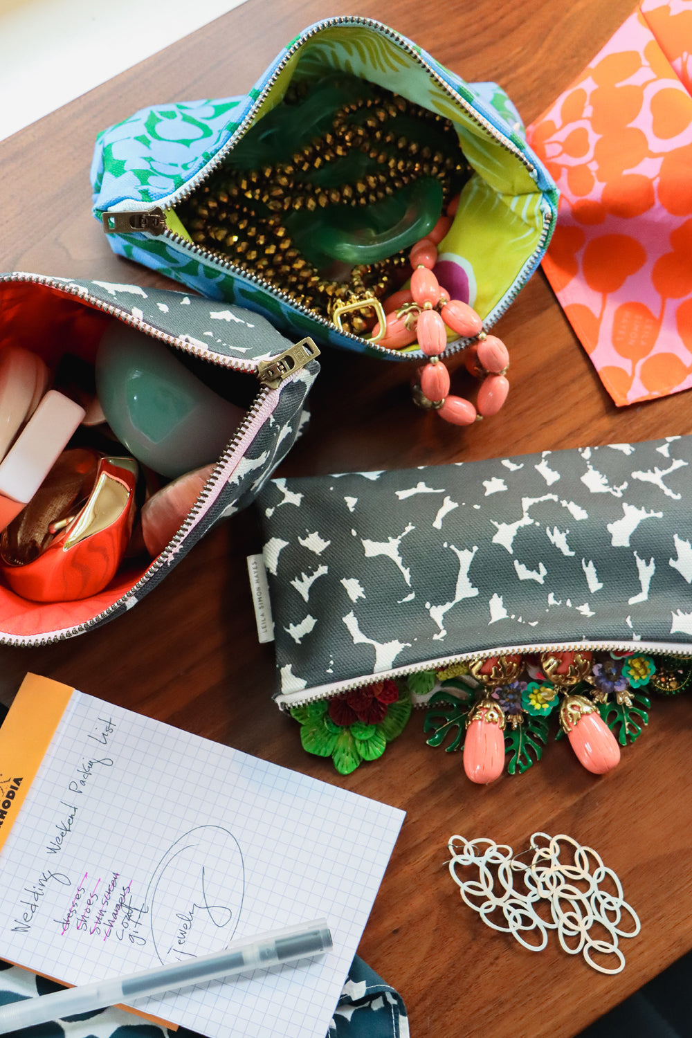 Three patterned zippered cosmetic pouches arranged on a wooden tabletop with jewelry and stationery props. The top pouch has a turquoise and green abstract print with a lime interior; the left pouch is dark gray with white abstract shapes and a coral interior; the right pouch is dark gray with white abstract shapes and a green floral trim.