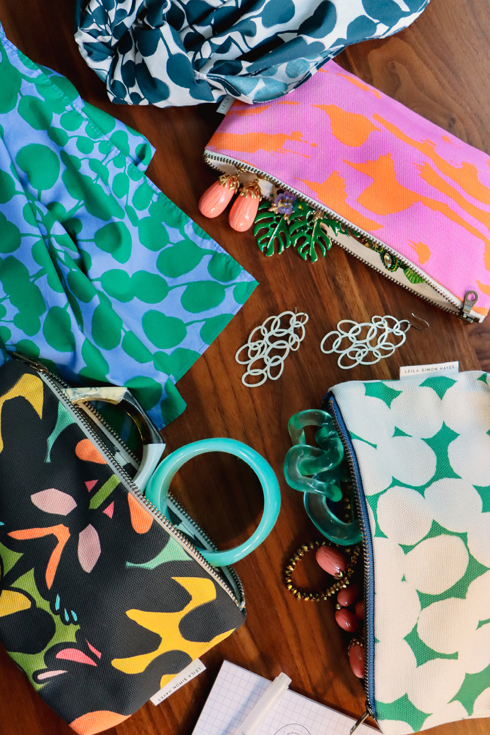 A styled flat-lay of several colorful patterned zip pouches and fabric accessories arranged on a wooden surface, with small jewelry pieces and papers nearby.