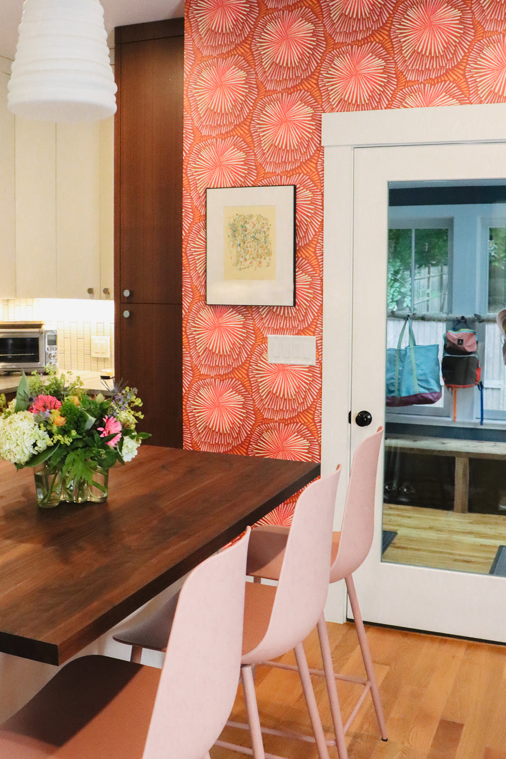 A dining area corner featuring a dark wood table and three light pink chairs, set against a wall with bright orange and pink patterned wallpaper. 