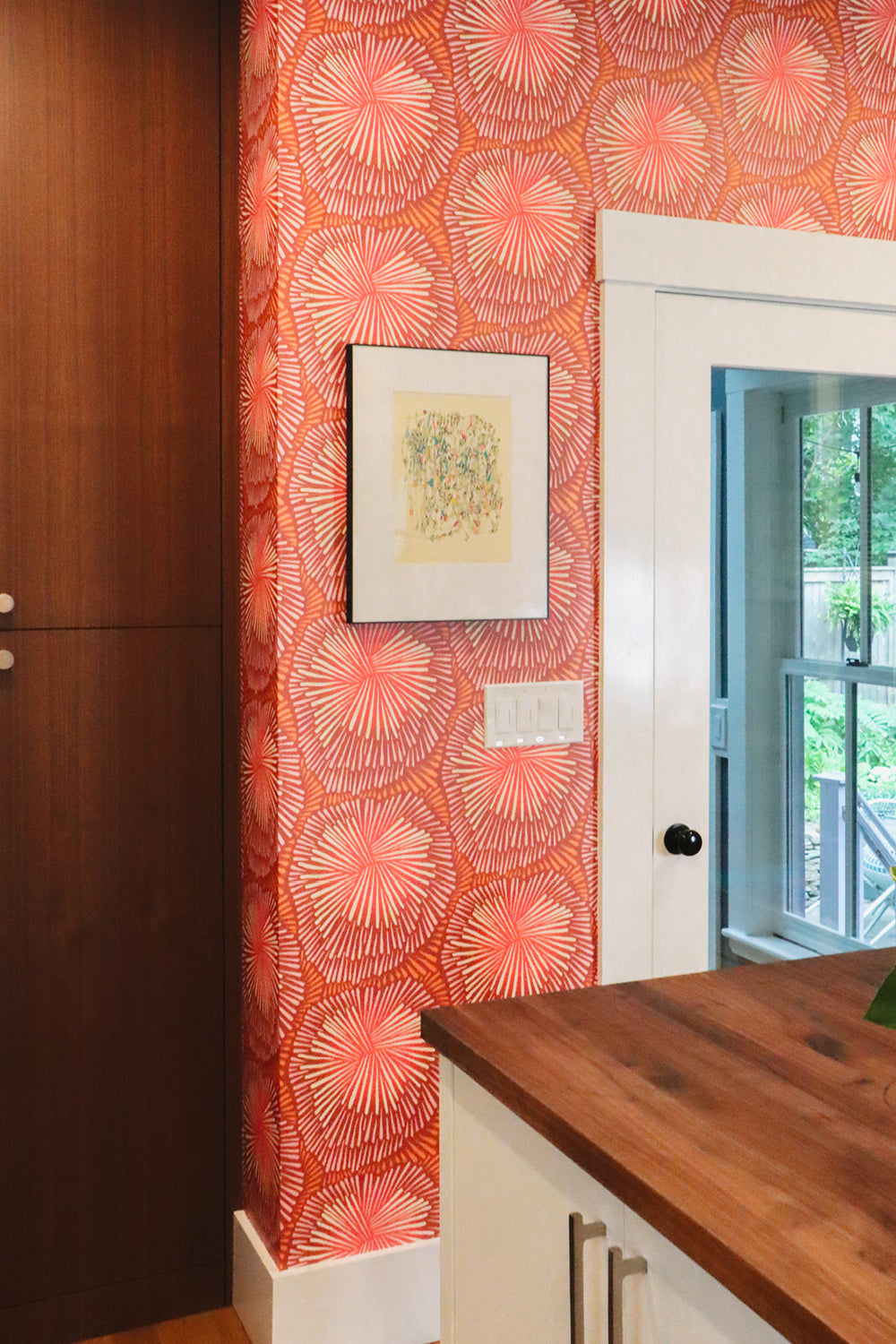 A close-up of a kitchen interior featuring a prominent wall covered in orange and pink fan-patterned wallpaper. A white door frame, a wooden countertop, and a dark wood cabinet are visible alongside the patterned wall.