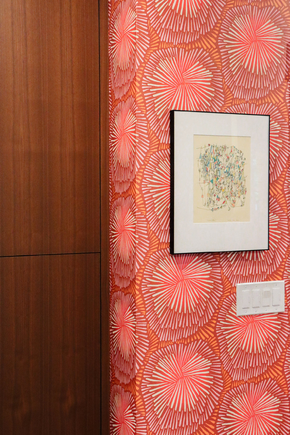 A close-up of a wall featuring bold coral and orange wallpaper with a repeating sunburst pattern, next to a dark wood cabinet. A small framed abstract print and a white wall switch are mounted on the wall.