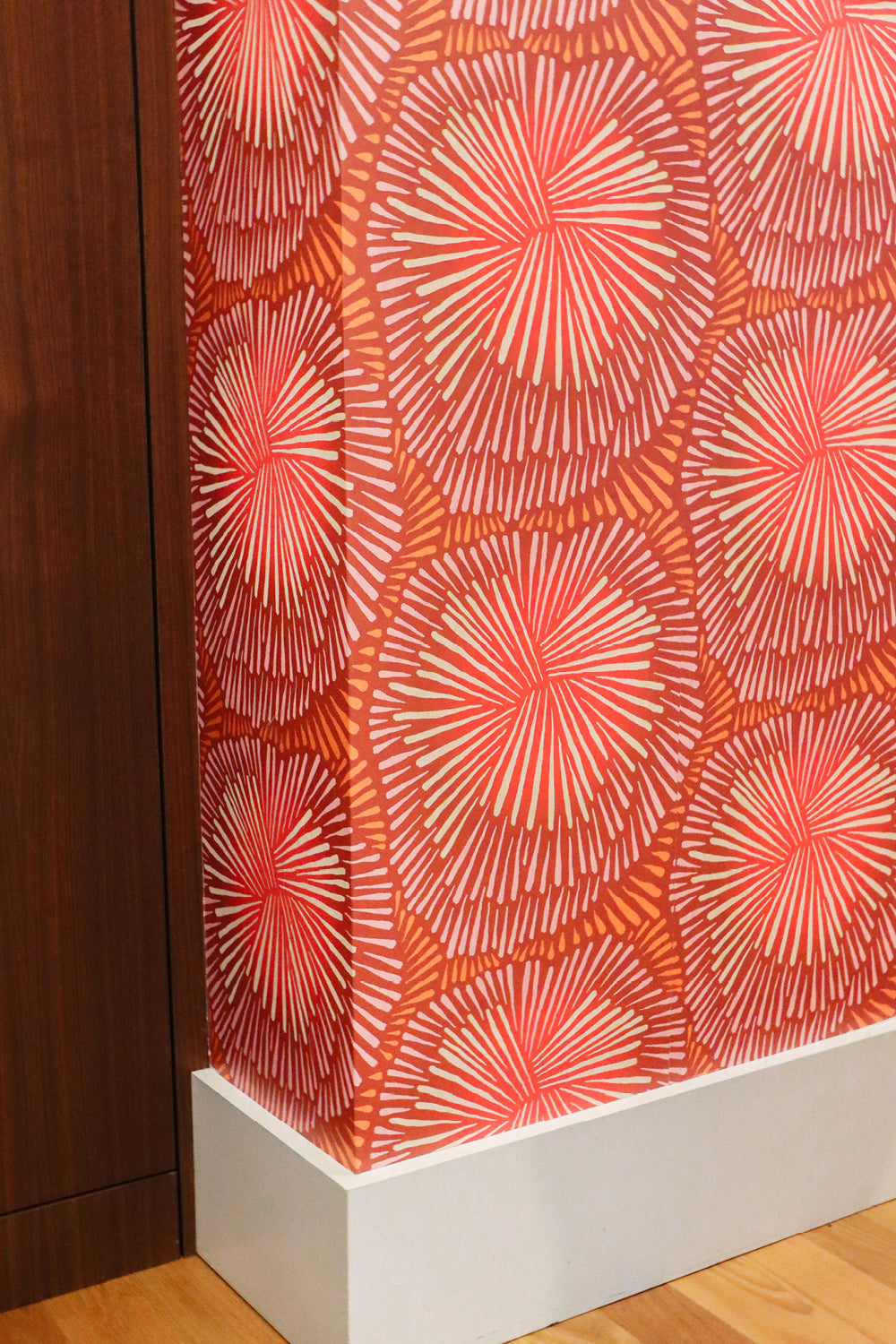 A close-up of wallpaper with a repeating coral and orange radial starburst pattern, shown installed on a wall above a white baseboard and light wood floor.