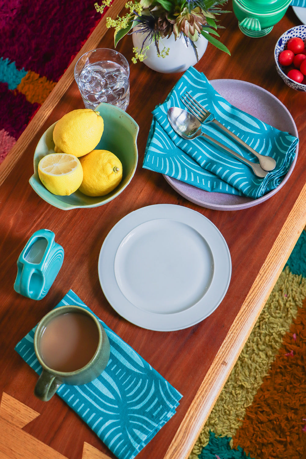 A styled tabletop scene with teal patterned napkins, a light blue plate, a green mug of coffee, lemons in a bowl, and other dining accessories arranged on a wooden table. Designed by artist Leila Simon Hayes.
