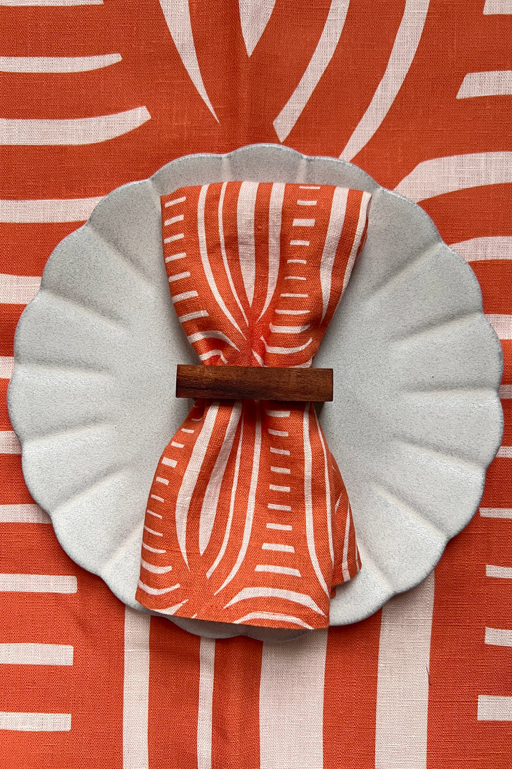 A coral colored linen napkin with a geometric pattern is displayed on a plate, over a table cloth with the same color and pattern. Designed by artist Leila Simon Hayes