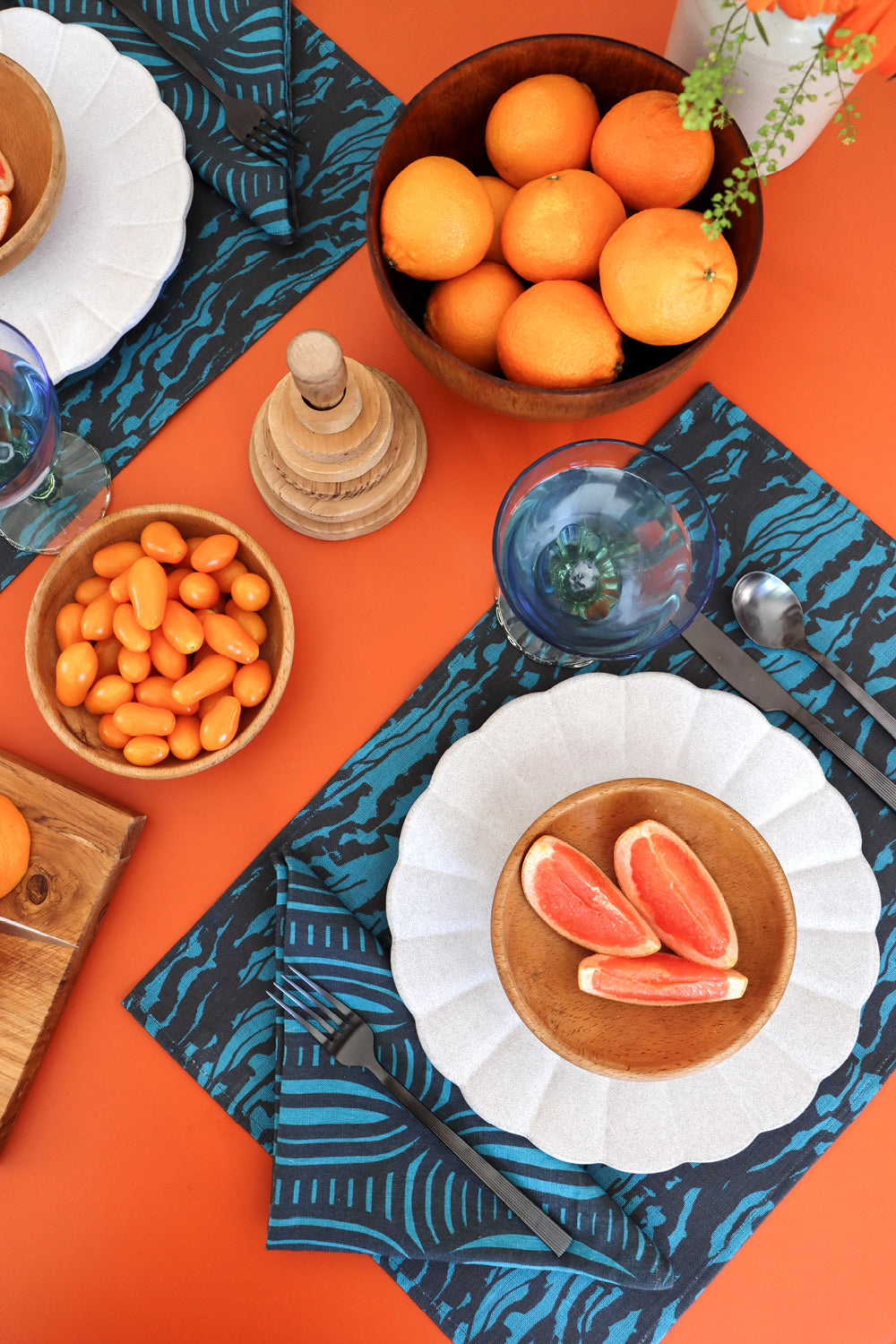 A blue linen placemat with a wave pattern, with a blue geometric napkin on an orange dining table with fruits. Designed by artist Leila Simon Hayes