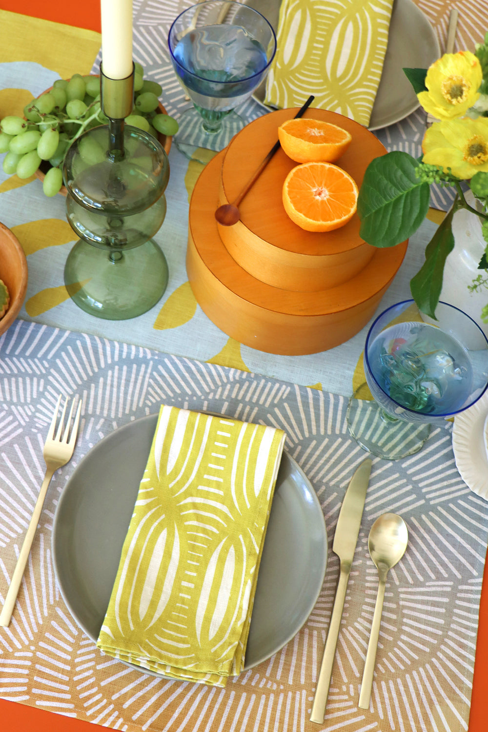 Colorful table setting with yellow patterned napkins, light blue and yellow gradient placemats with an organic line pattern, and cut fruit on a geometric table green runner. Designed by artist Leila Simon Hayes