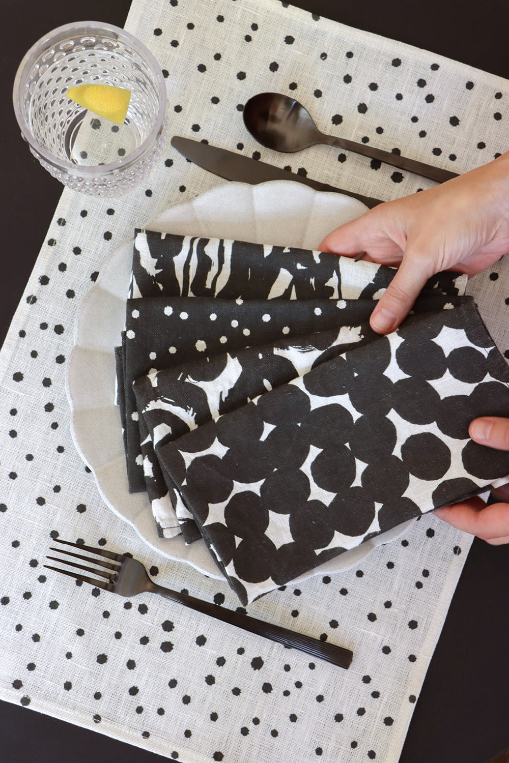 A set of four black and white linen napkins with a geometric pattern being held over a set table with a white placemat with small dots. Designed by artist Leila Simon Hayes.