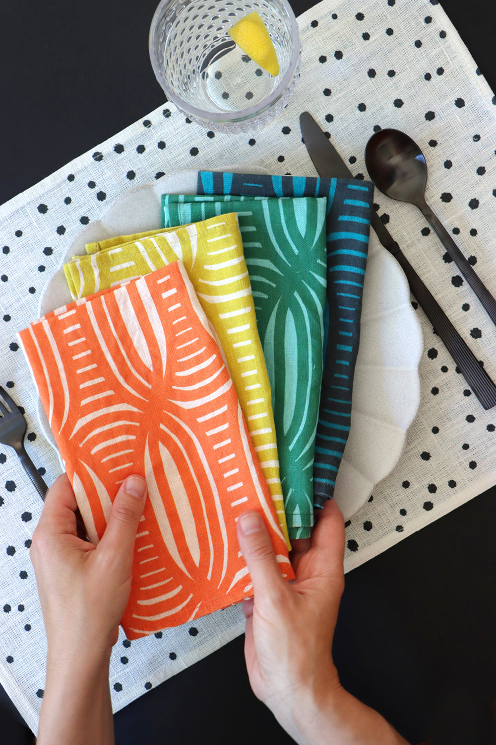 Colorful patterned linen napkins held by hands on a black and white polka dot tablecloth with cutlery. Designed by artist Leila Simon Hayes