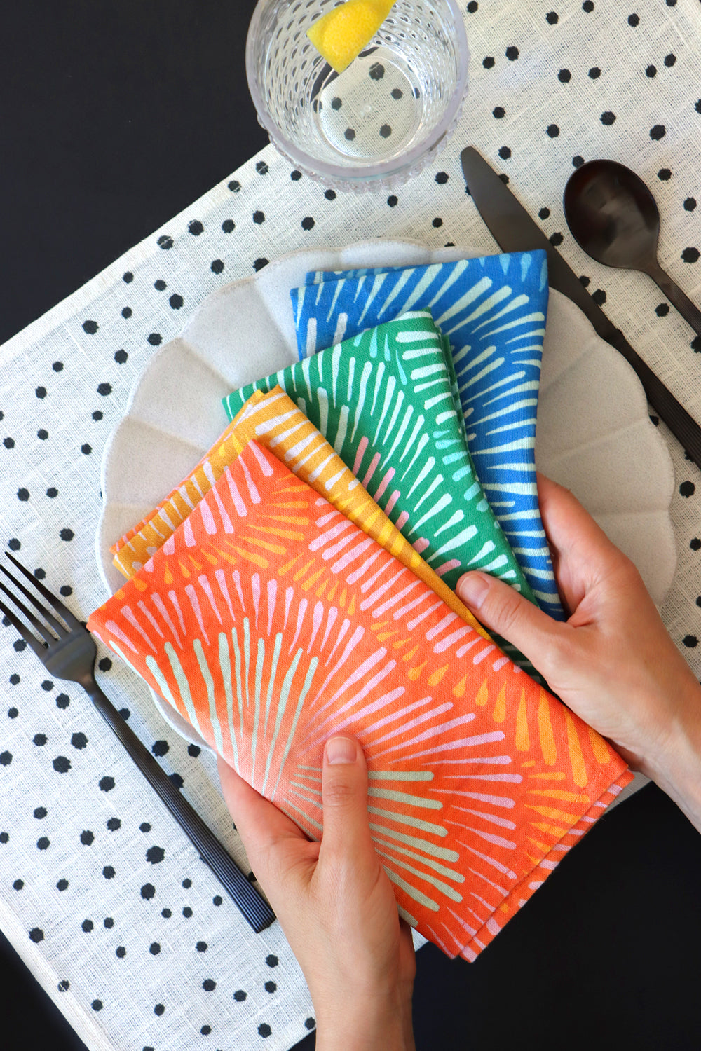 Four colorful patterned napkins being held over a plate with cutlery on a black and white polka dot placemat.