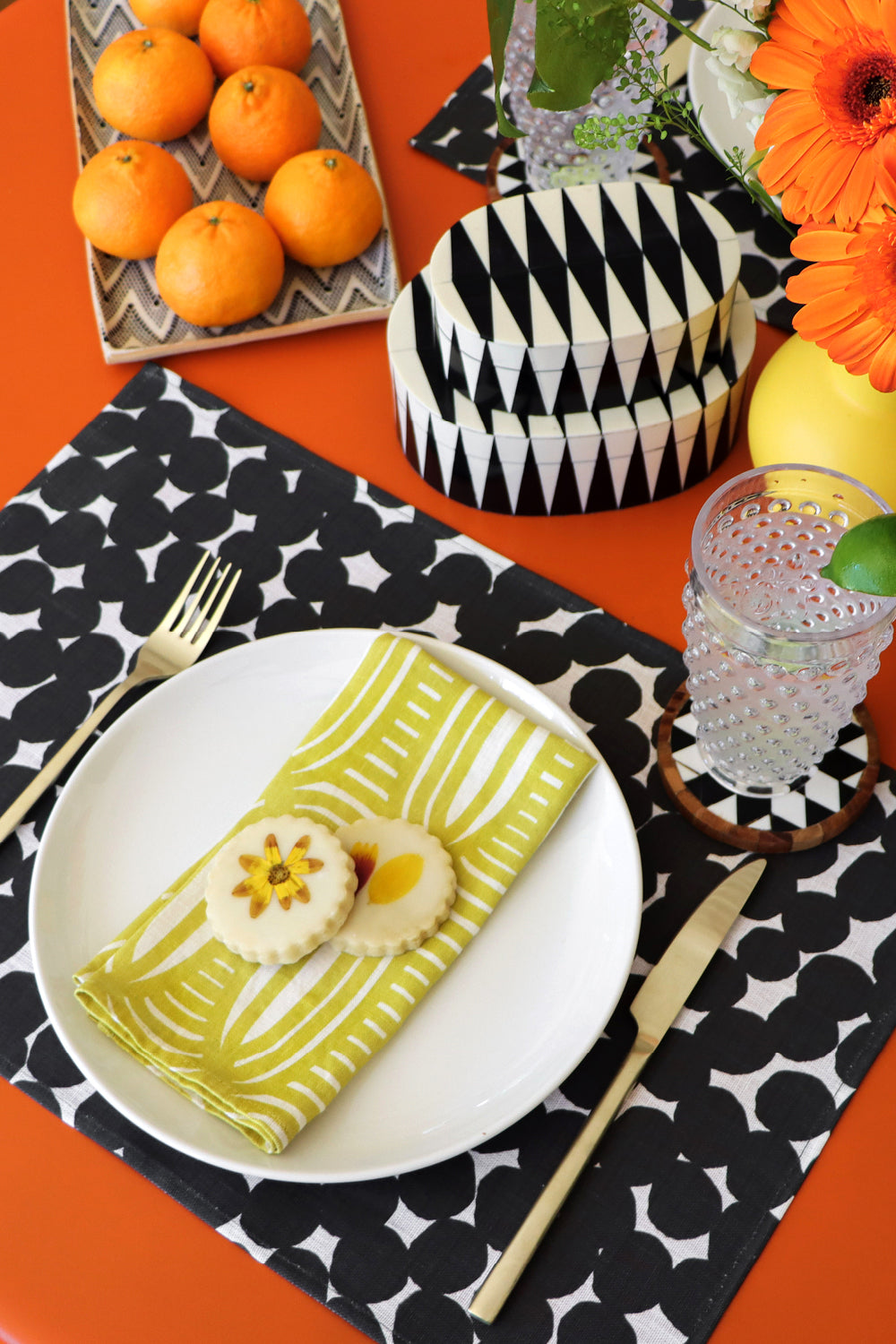 Dinner setup with a plate of food, cutlery, and decorative elements on a black and white dots patterned placemat and yellow geometric striped linen napkin. Designed by artist Leila Simon Hayes