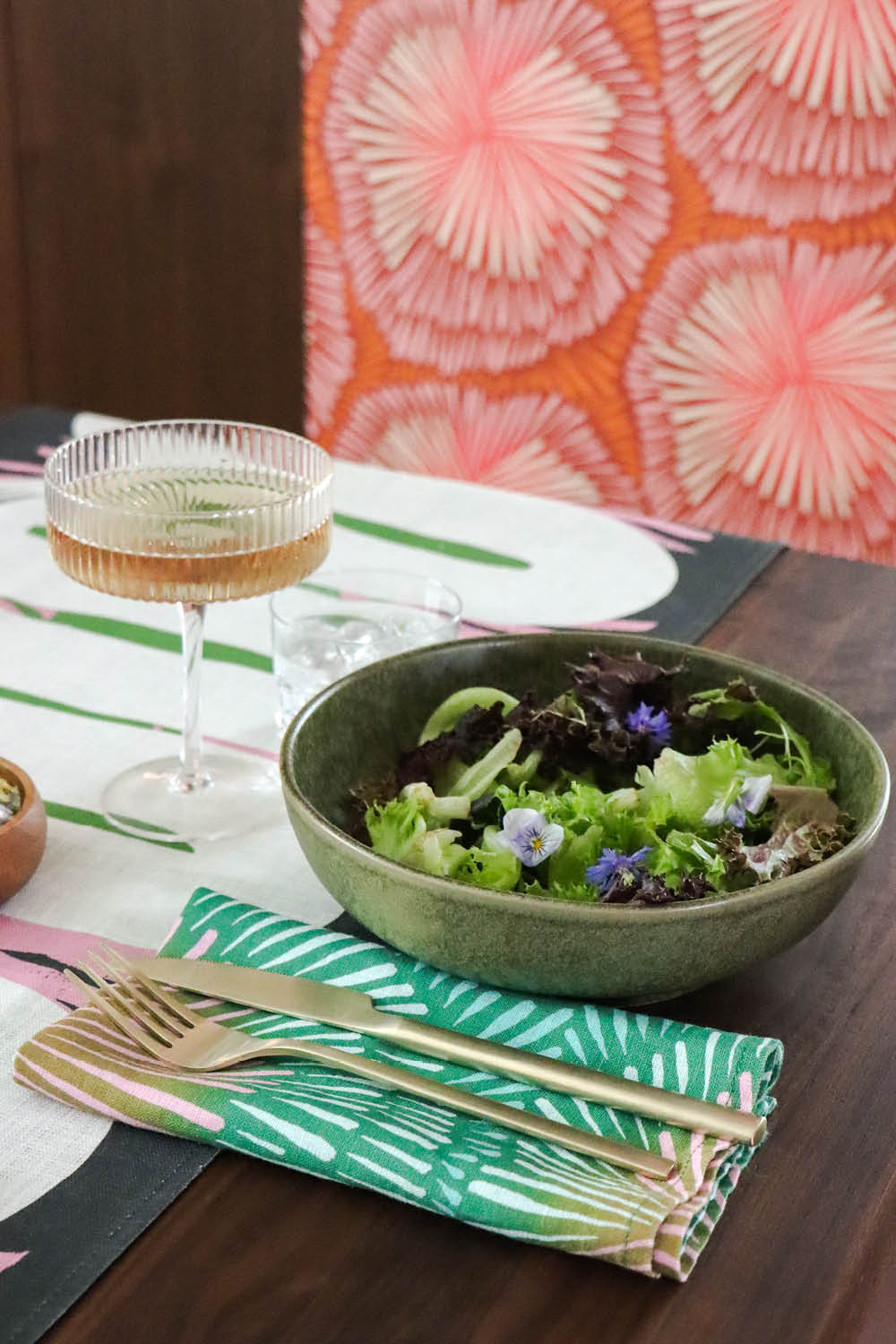 Dining table setting with a bowl of salad, glass, and cutlery on a green burst napkin and abstract table runner. Designed by artist Leila Simon Hayes. 