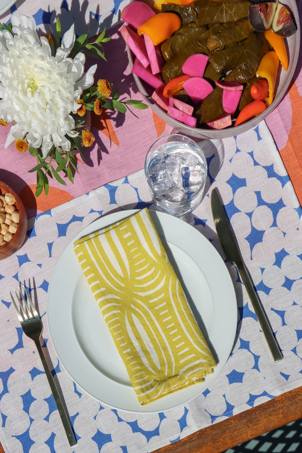 Colorful table setting with a plate, cutlery, and yellow napkin on a blue and white dotted patterned placemat over a pink and coral table runner. Designed by artist Leila Simon Hayes