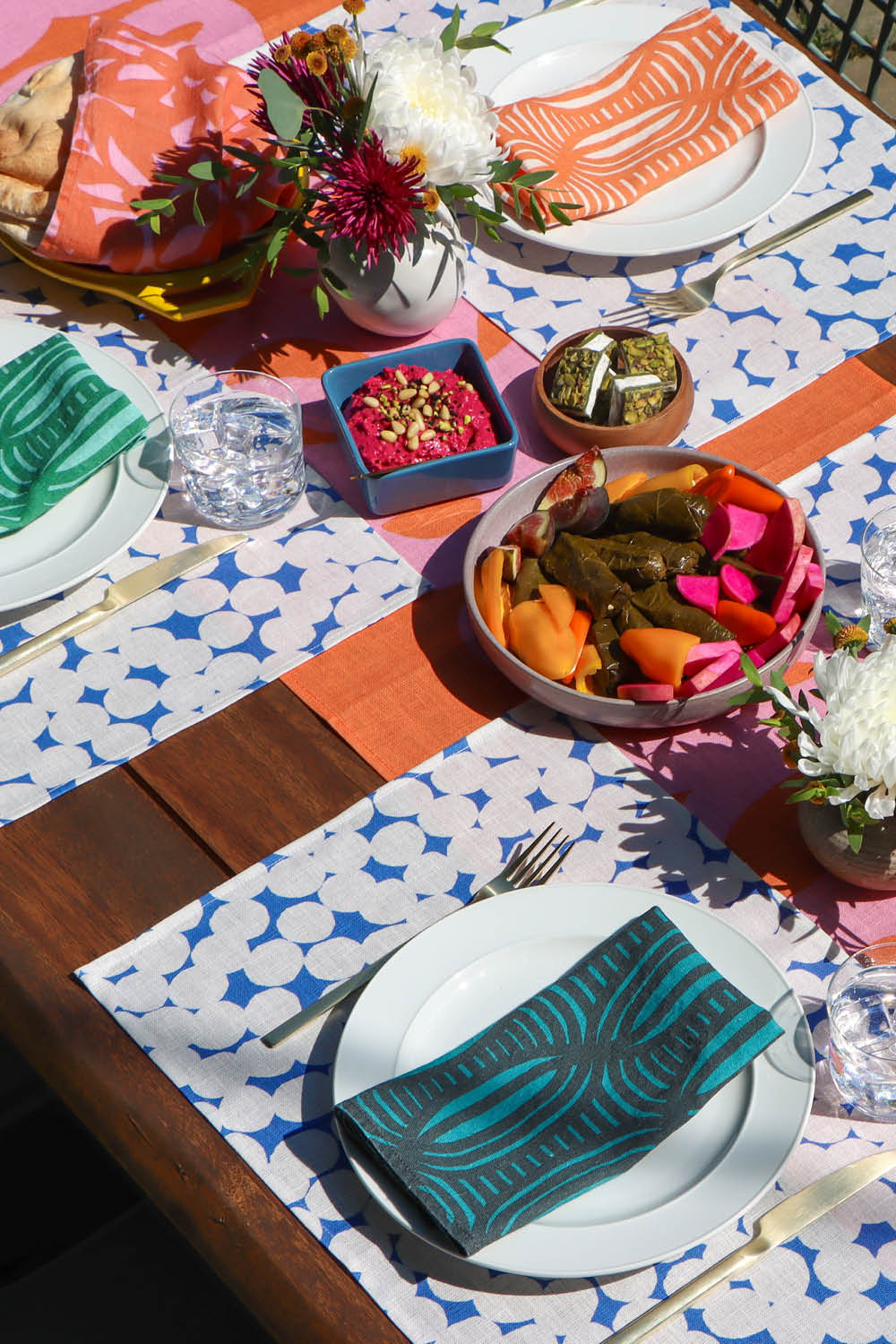 Colorful table setting with blue and white dots patterned placemats, colorful striped napkins, and food on a coral geometric table runner. Designed by artist Leila Simon Hayes
