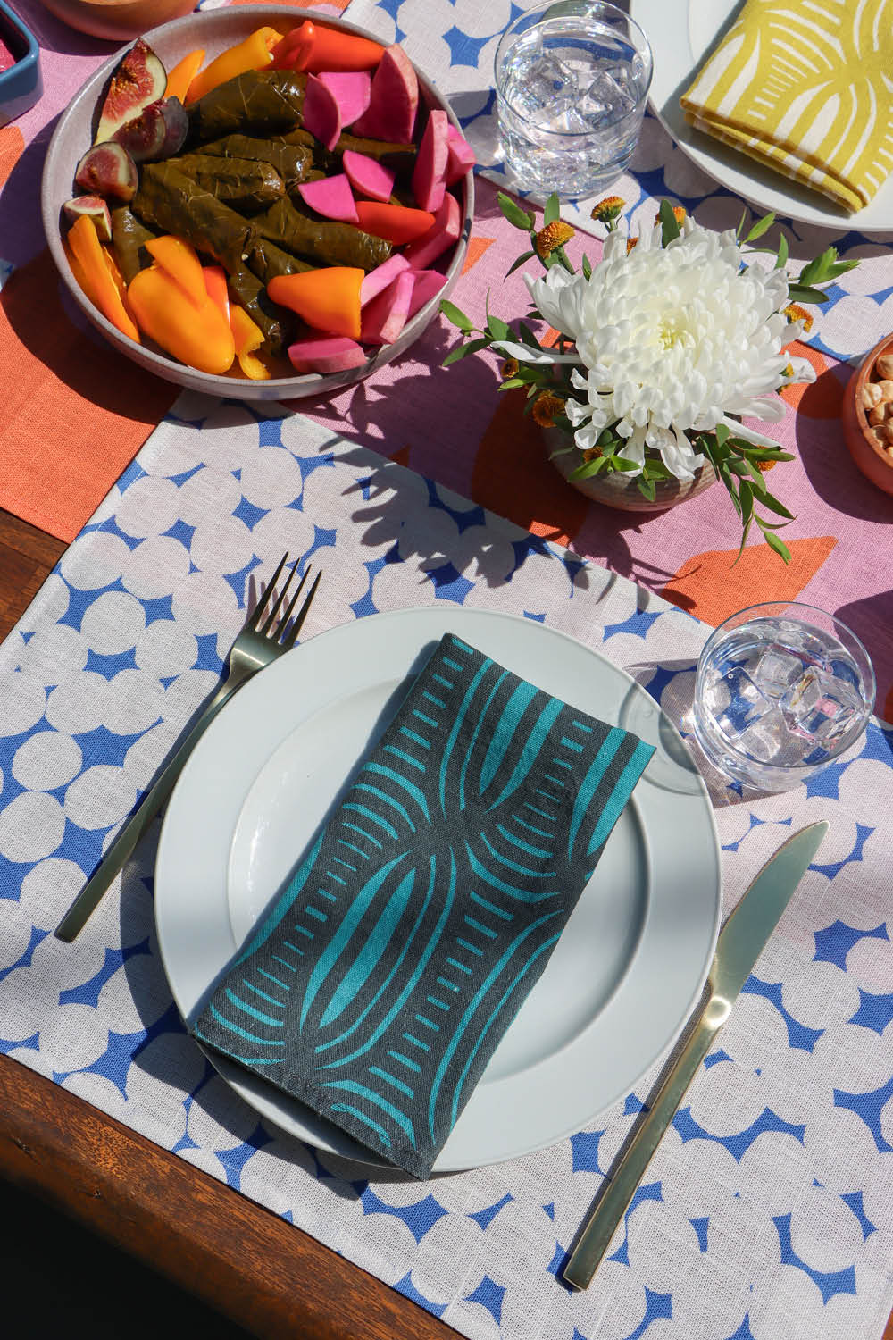 Dining table setting with a colorful striped dinner napkins, blue and white dots placemats, over a pink and coral table runner. Designed by artist Leila Simon Hayes