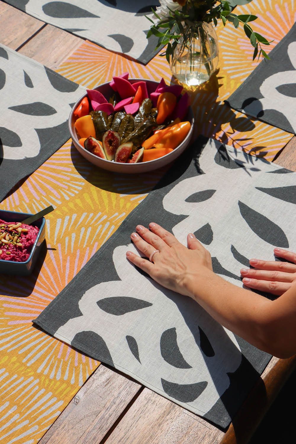Hands setting up a colorful outdoor setting. Black and white abstract placemats, and an orange and yellow burst patterned table runner, designed by artist Leila Simon Hayes.