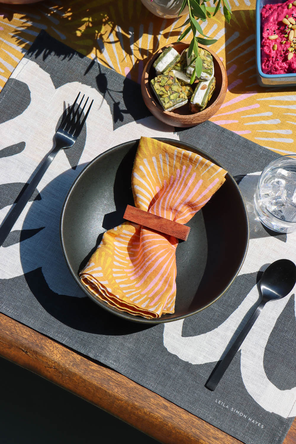 Dining table setting with an orange sun burst napkin on a black plate, over a black and white abstract placemat, over a table runner with the same napkin pattern. Designed by artist Leila Simon Hayes. 