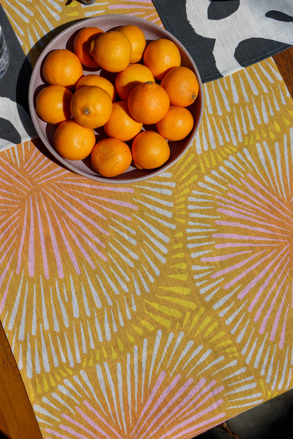 A close up of the table runner design showing the yellow and orange tones and burst pattern on a table next to black and white placemats and a bowl of oranges. Designed by artist Leila Simon Hayes.