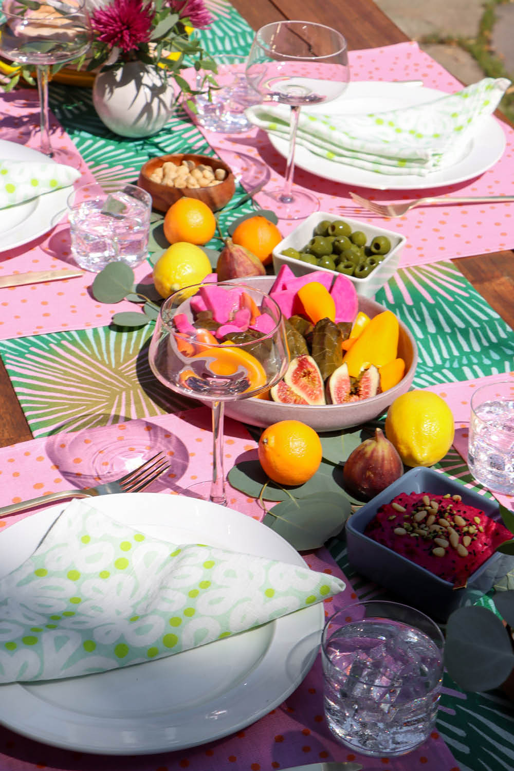 Colorful outdoor dining table setting with green abstract napkins, pink polka dot placemats, and a green burst patterned table runner. Designed by artist Leila Simon Hayes.