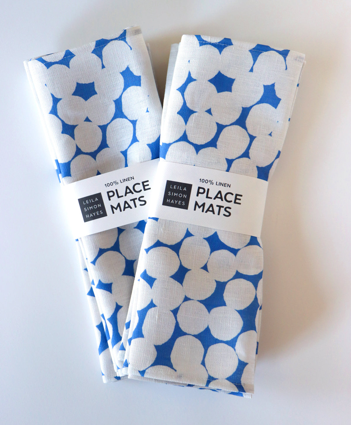 Four blue and white patterned place mats with '100% linen Placemats' labels, designed by artist Leila Simon Hayes on a light background.