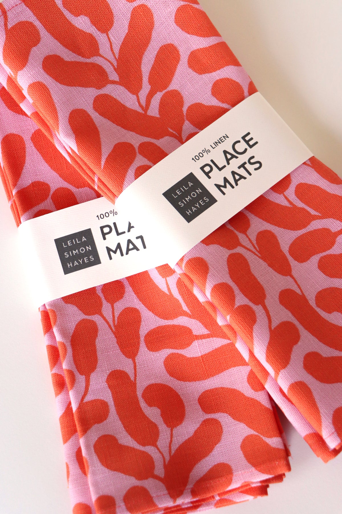 Four folded placemats with a pink background and coral botanical leaf design. Wrapped in a label saying "100% Linen Placemats", designed by artist Leila Simon Hayes