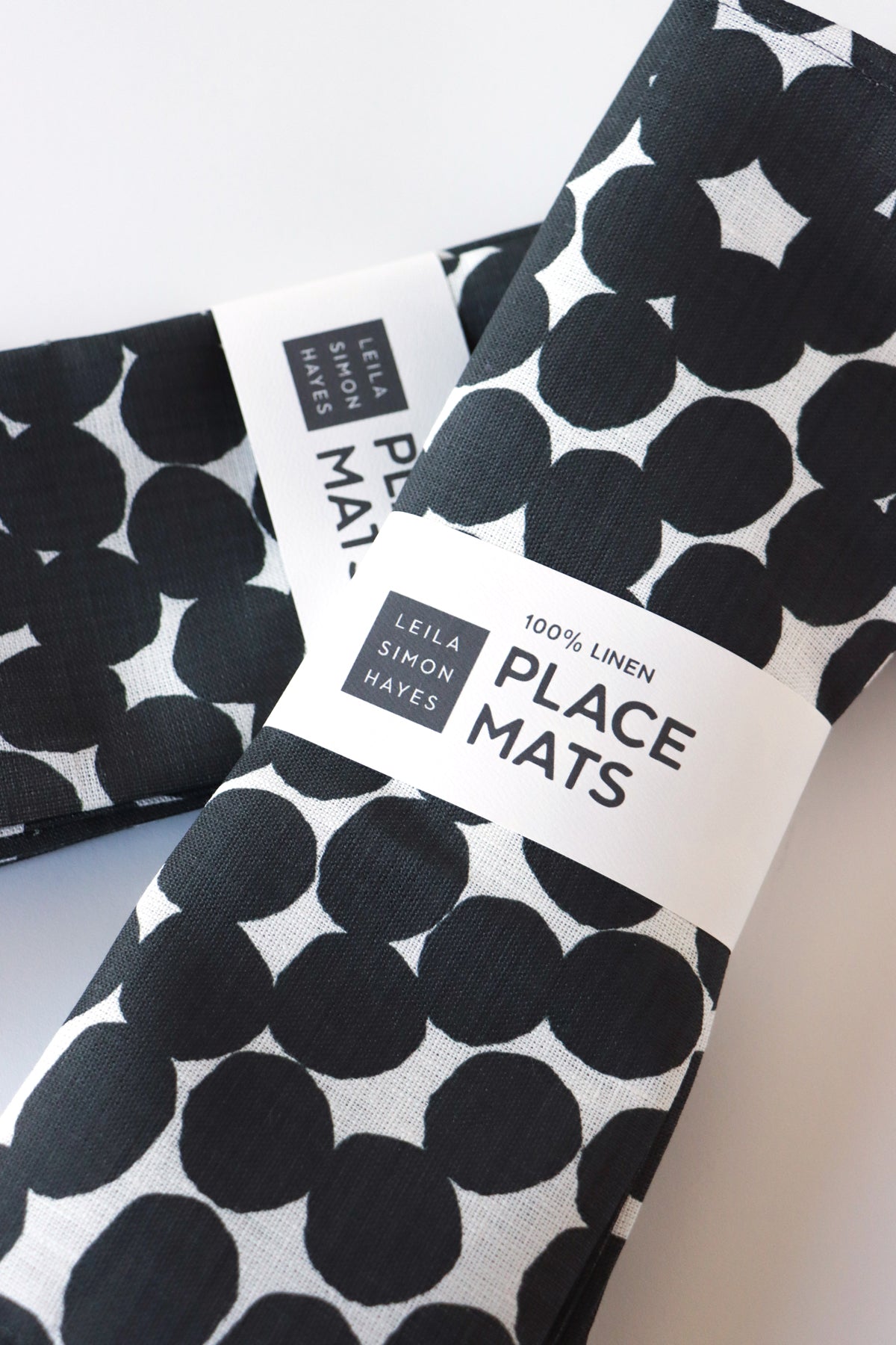 Set of four, black and white dots patterned placemats with brand labels saying "100% Linen Placemats" on a light background. Designed by artist Leila Simon Hayes