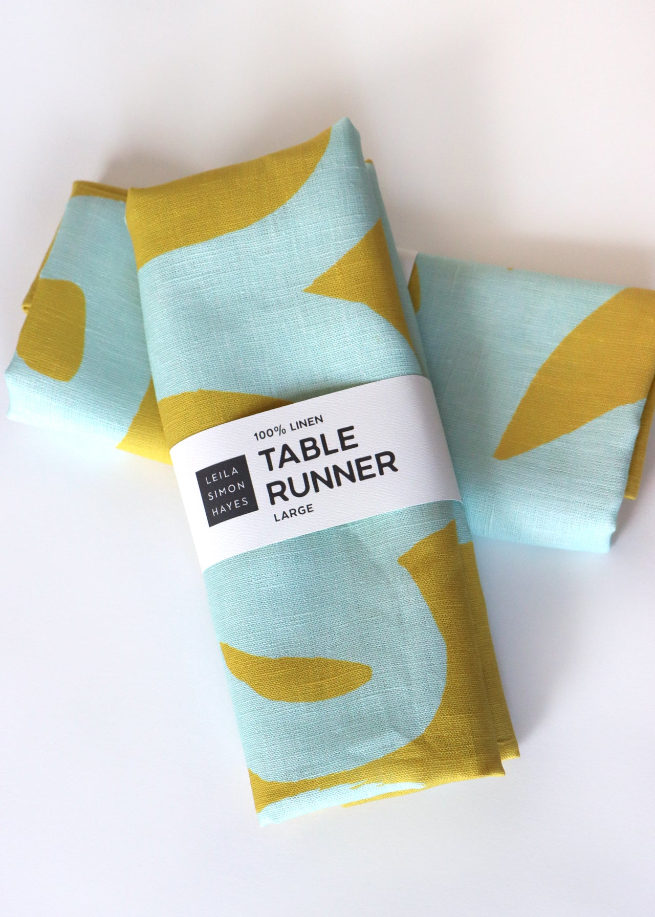 Packaged table runner with a label saying "100% Linen Table Runner", designed by artist Leila Simon Hayes.