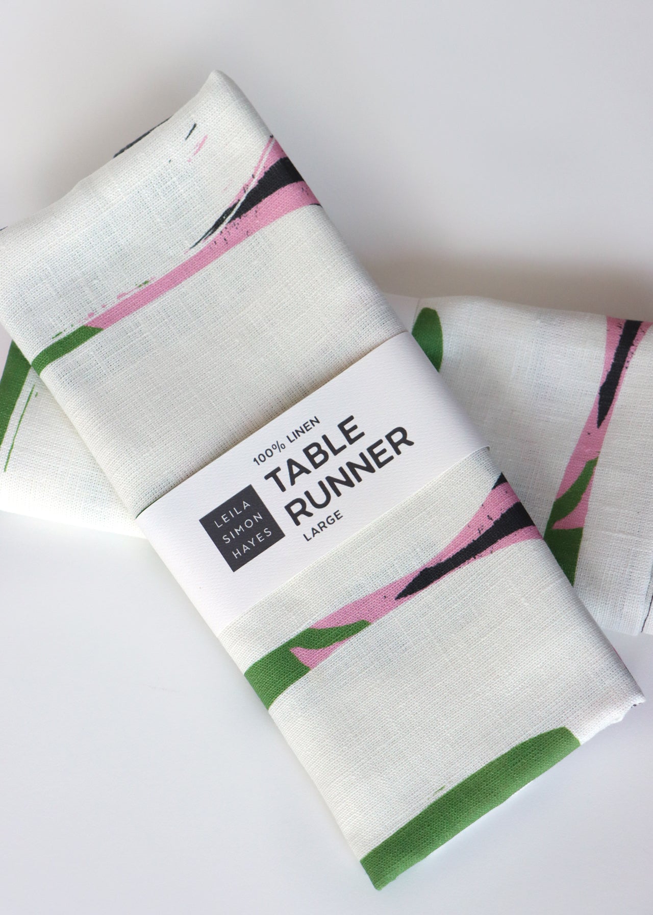 Packaged table runner with pink and green design on a white background. The label says "100% Linen Table Runner", designed by artist Leila Simon Hayes.