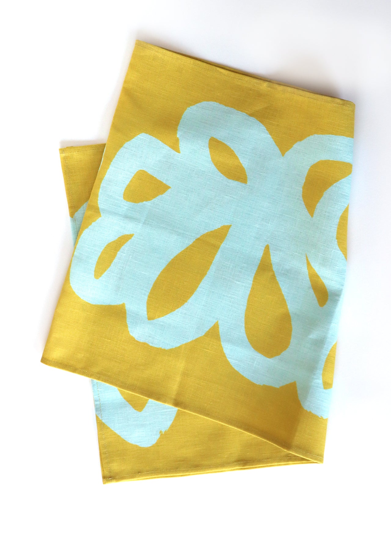 A green table runner with a light blue floral lace pattern, designed by artist Leila Simon Hayes. The table runner is folded over a white background. 