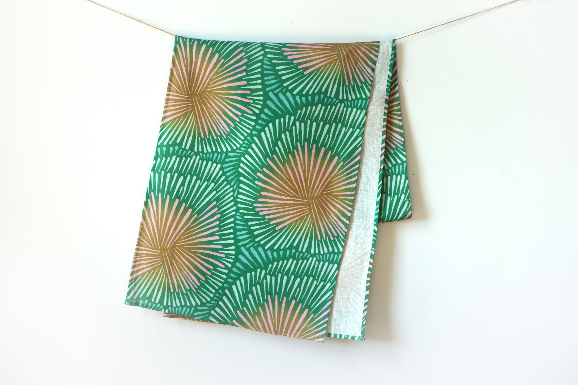 A green table runner with pink accents in a burst pattern hanging on a white background, designed by artist Leila Simon Hayes.