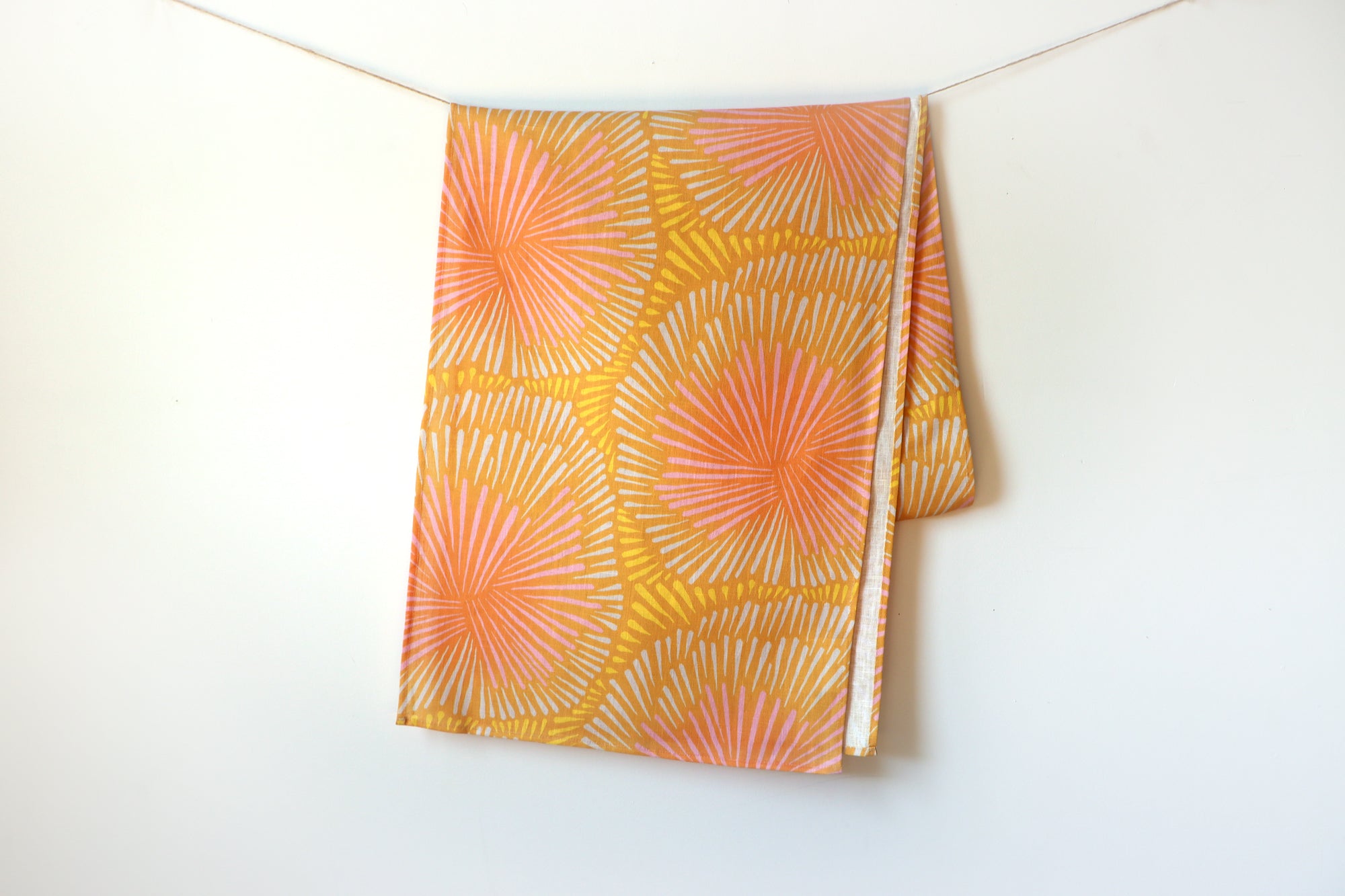 Orange and yellow table runner with a burst pattern hanging on a white background, designed by artist Leila Simon Hayes.