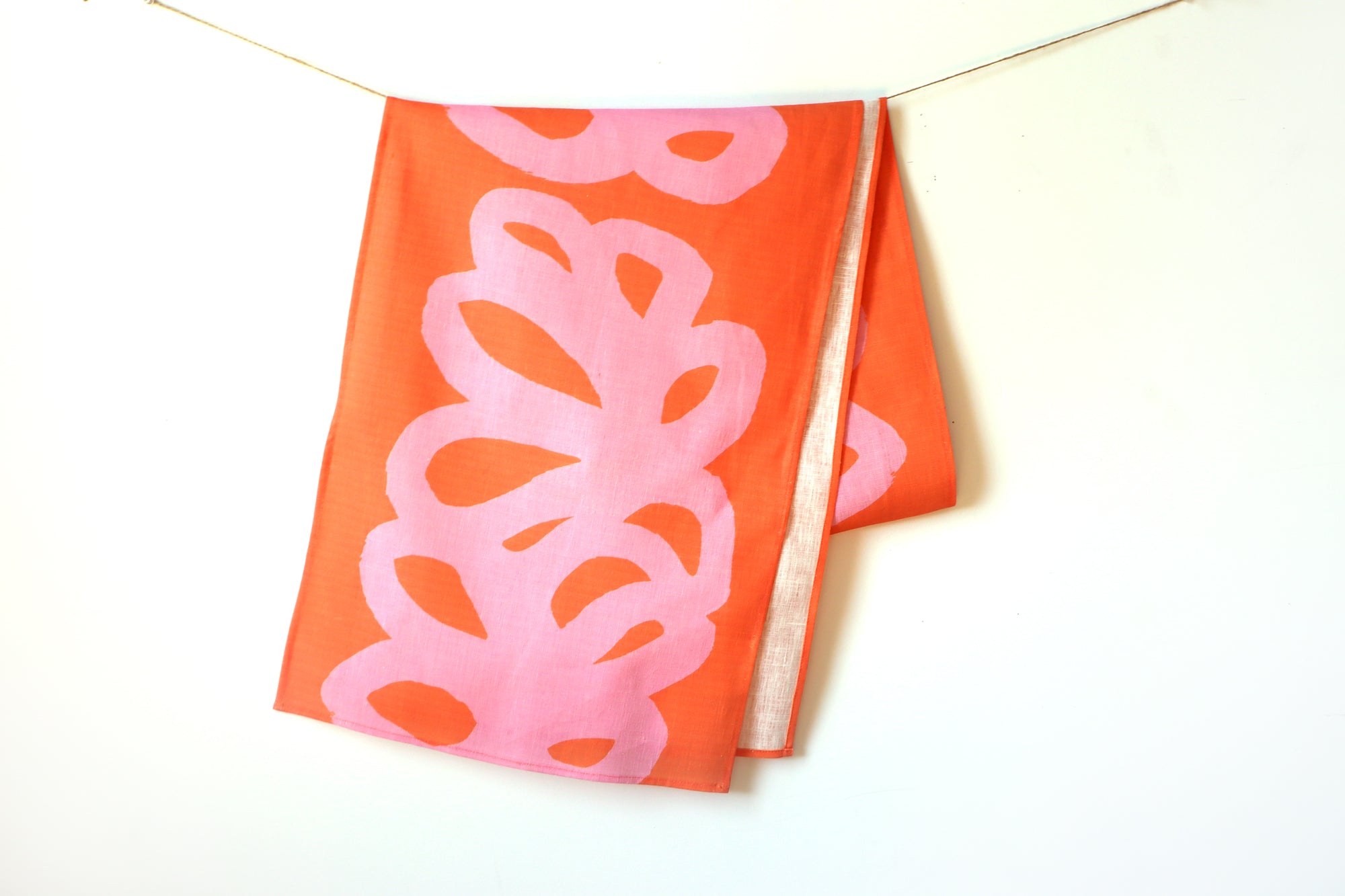 A coral table runner with a pink abstract pattern hanging on a white background, designed by artist Leila Simon Hayes.