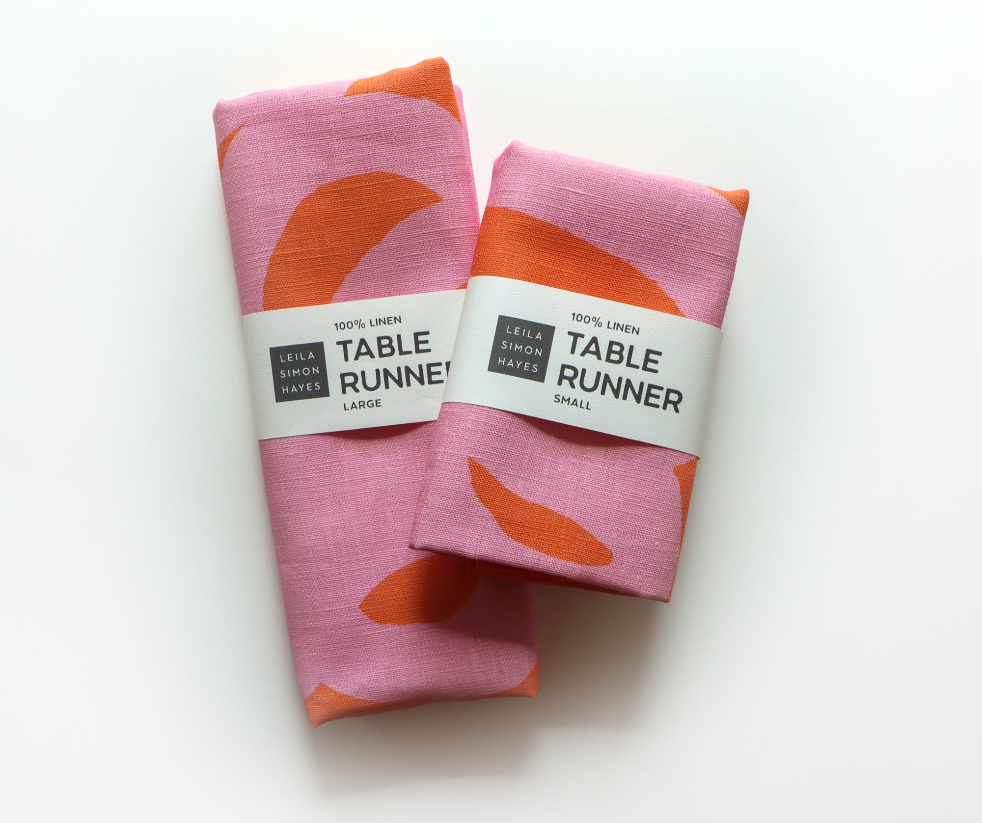 Two coral table runners with pink abstract patterns and labels saying "100% Linen Table Runner", designed by artist Leila Simon Hayes.