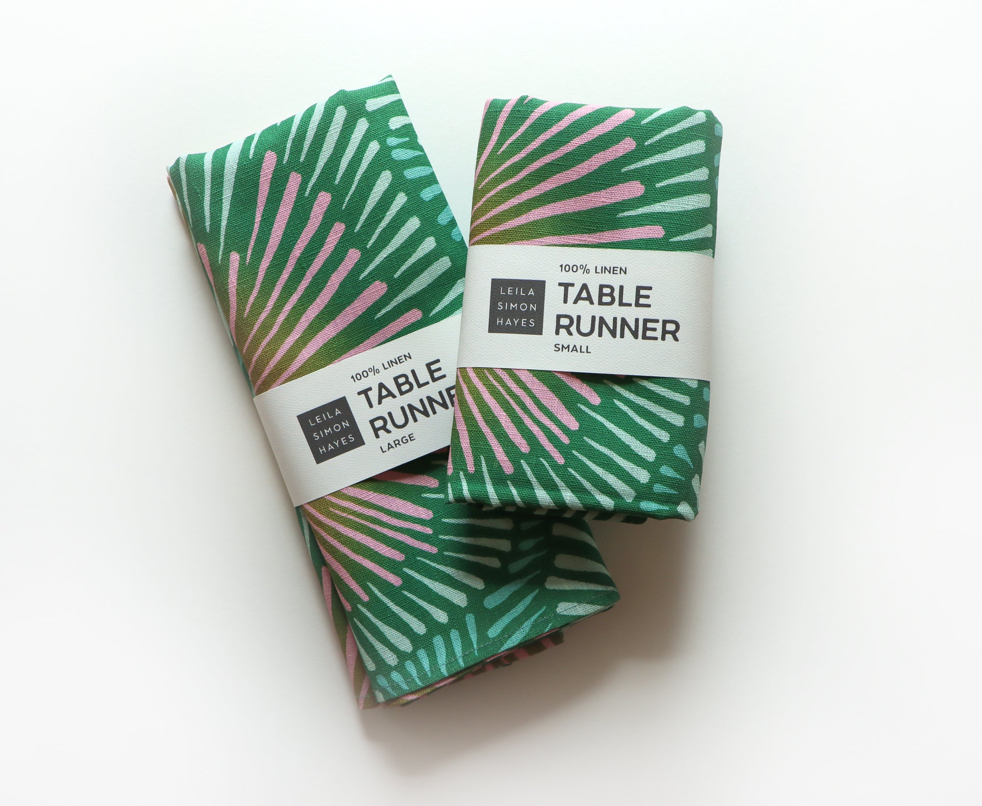 Two folded green table runners with pink accents in a burst pattern with labels saying "100% Linen Table Runner", designed by artist Leila Simon Hayes.