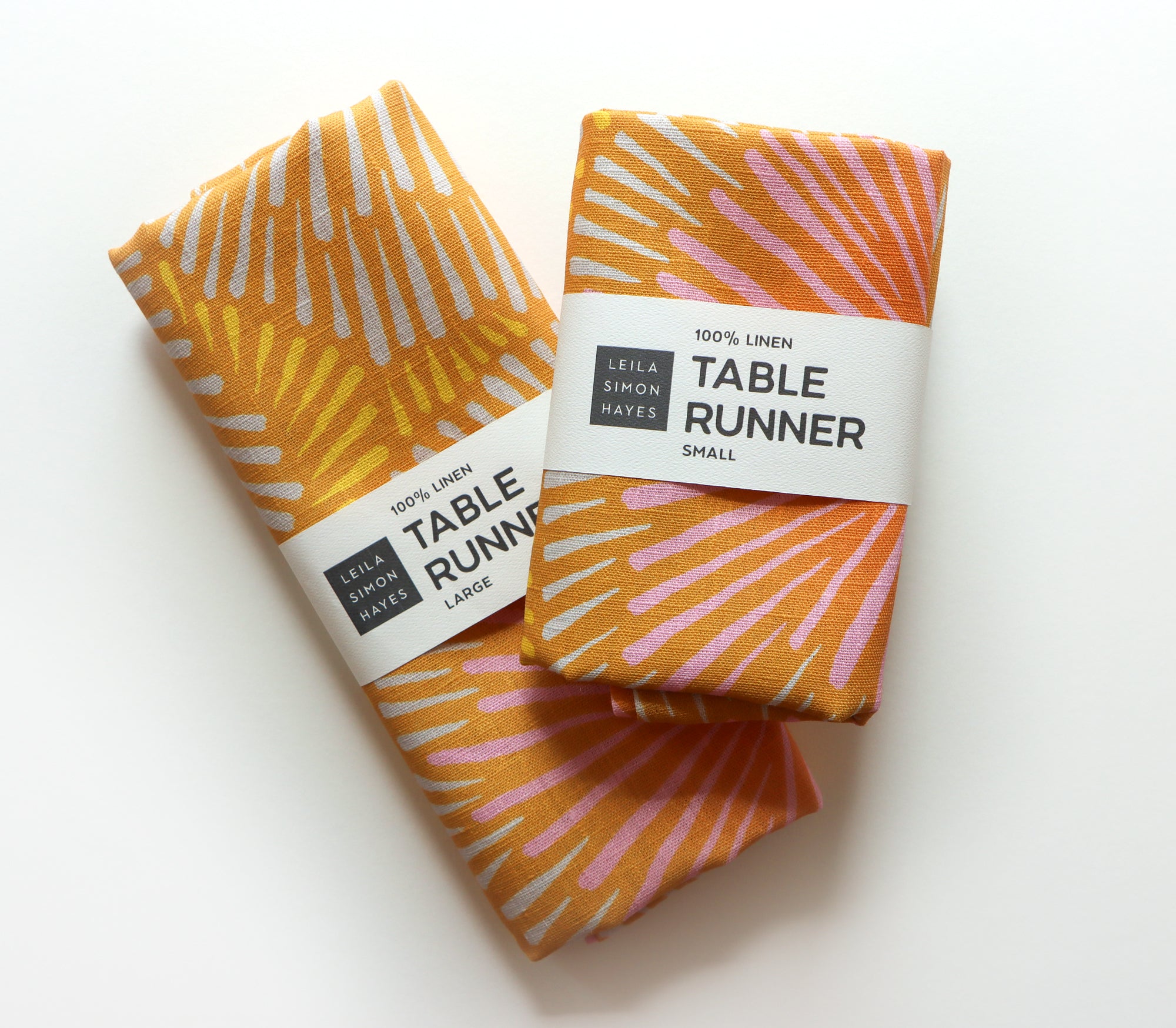 Two folded yellow and orange table runners with a burst pattern. They each have labels saying "100% Linen Table Runner", designed by artist Leila Simon Hayes.