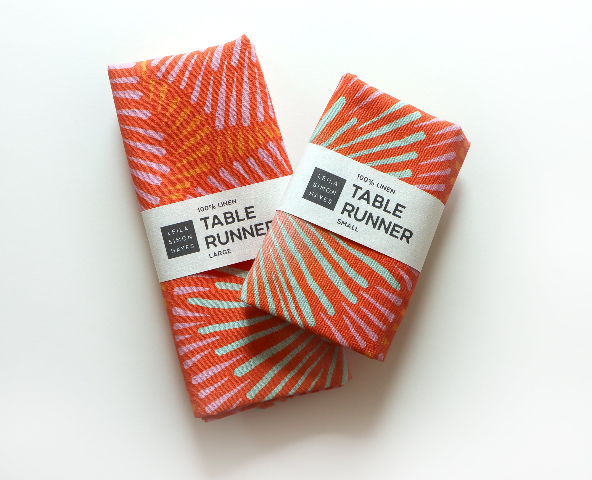 Two folded red table runners with a colorful burst pattern on a white background. Labels say "100% Linen Table Runner", designed by artist Leila Simon Hayes.