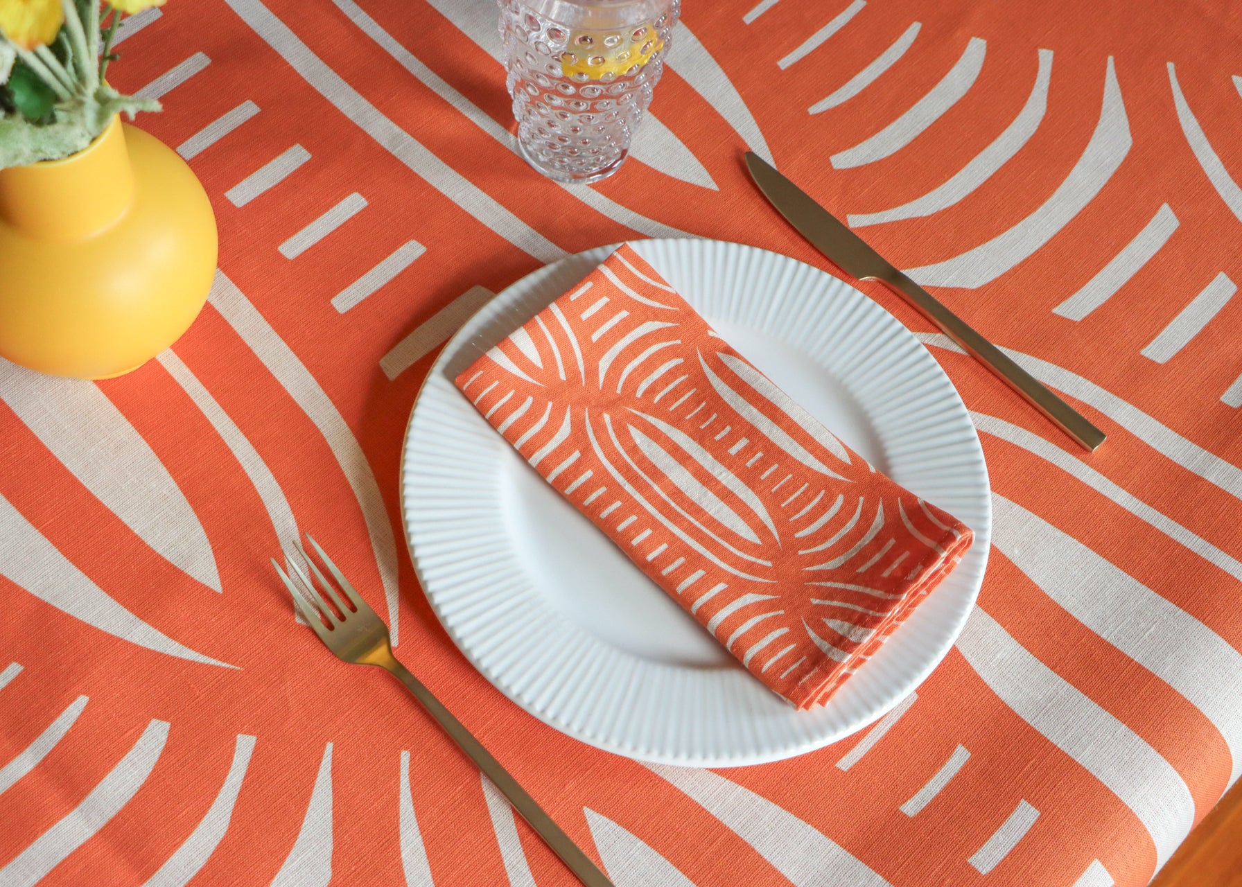 Table setting with a coral tablecloth that has a geometrical stripe pattern with a white plate and napkin with the design on top. Designed by artist Leila Simon Hayes.