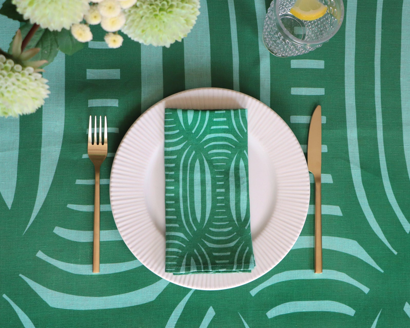 Table setting with a green patterned napkin on a white plate, gold fork and knife, and floral arrangement on a green geometric patterned tablecloth. 