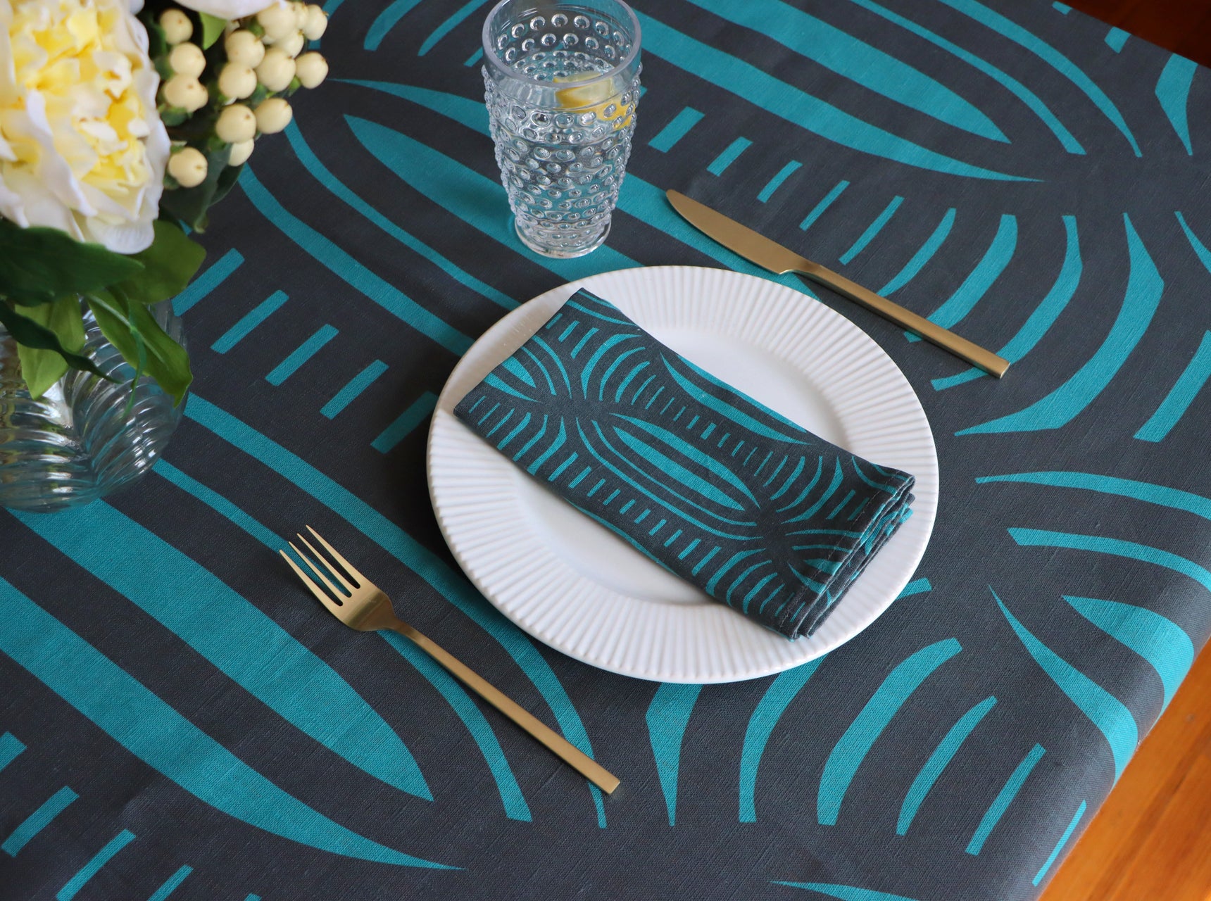 Table setting with a dark blue and teal patterned tablecloth, white plate, green napkin, and gold cutlery. Designed by artist Leila Simon Hayes.