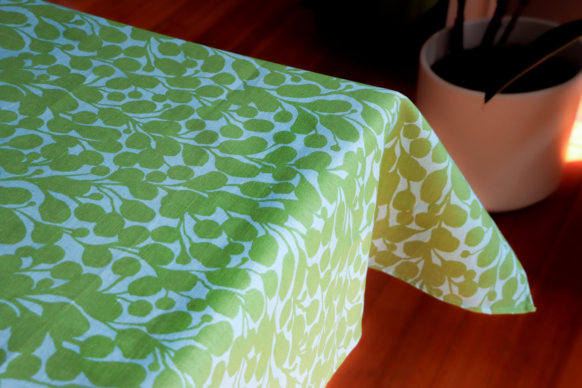 Green and blue floral patterned tablecloth on a wooden surface with a plant in the background. Designed by artist Leila Simon Hayes.