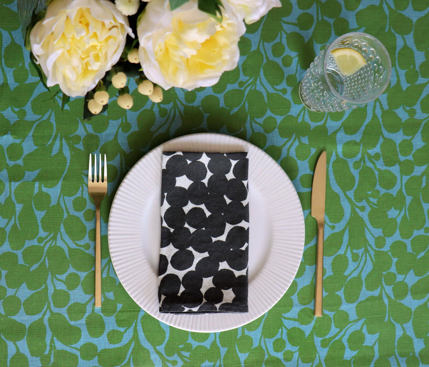 Table setting with a black and white dot patterned napkin on a white plate, gold fork and knife, and yellow flowers on a green and blue floral and botanical patterned tablecloth. Designed by artist Leila Simon Hayes.
