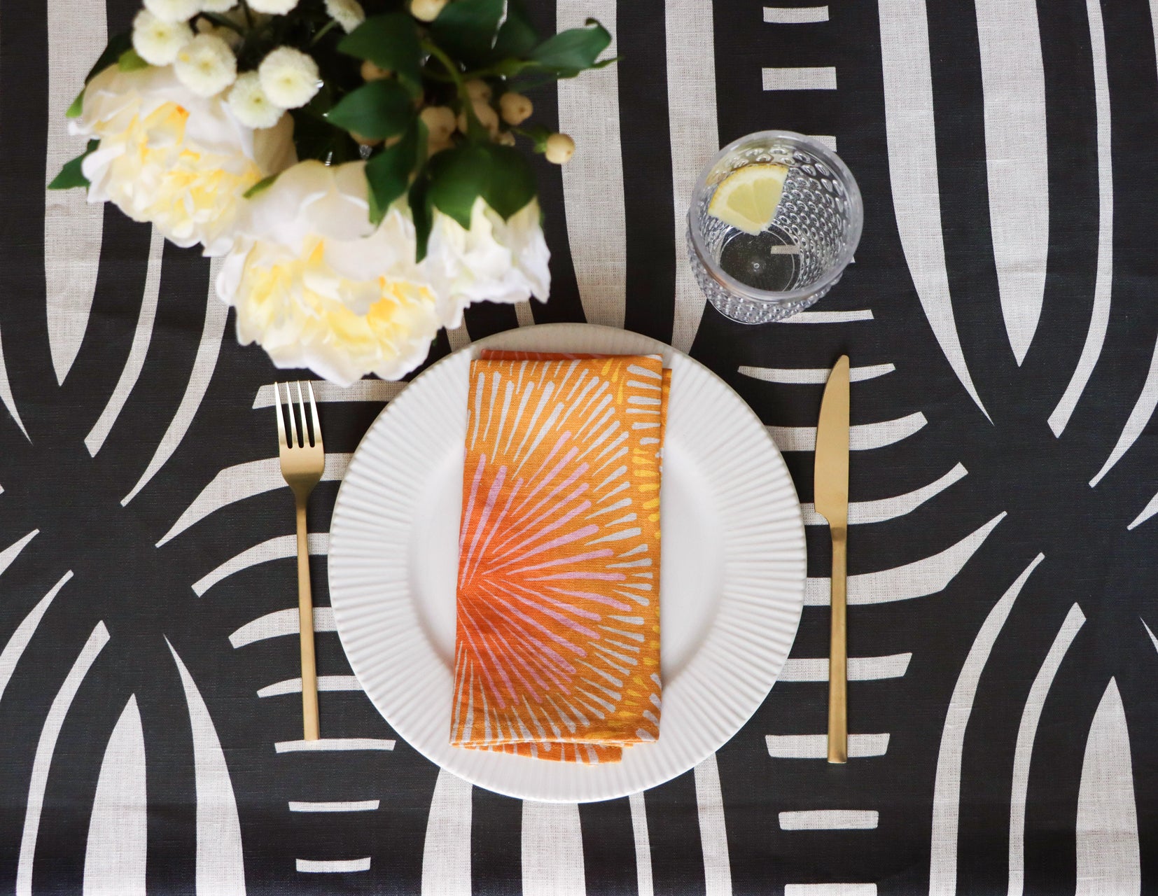 Dinner setting with a black and white patterned tablecloth, white plate with an orange napkin, gold fork and knife, and white flowers. Table linens designed by artist Leila Simon Hayes.