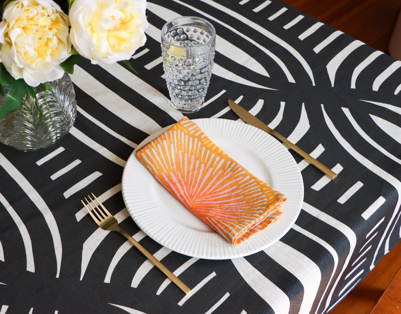 Folded orange burst napkin on a plate with a black and white striped table cloth behind it. Designed by artist Leila Simon Hayes.
