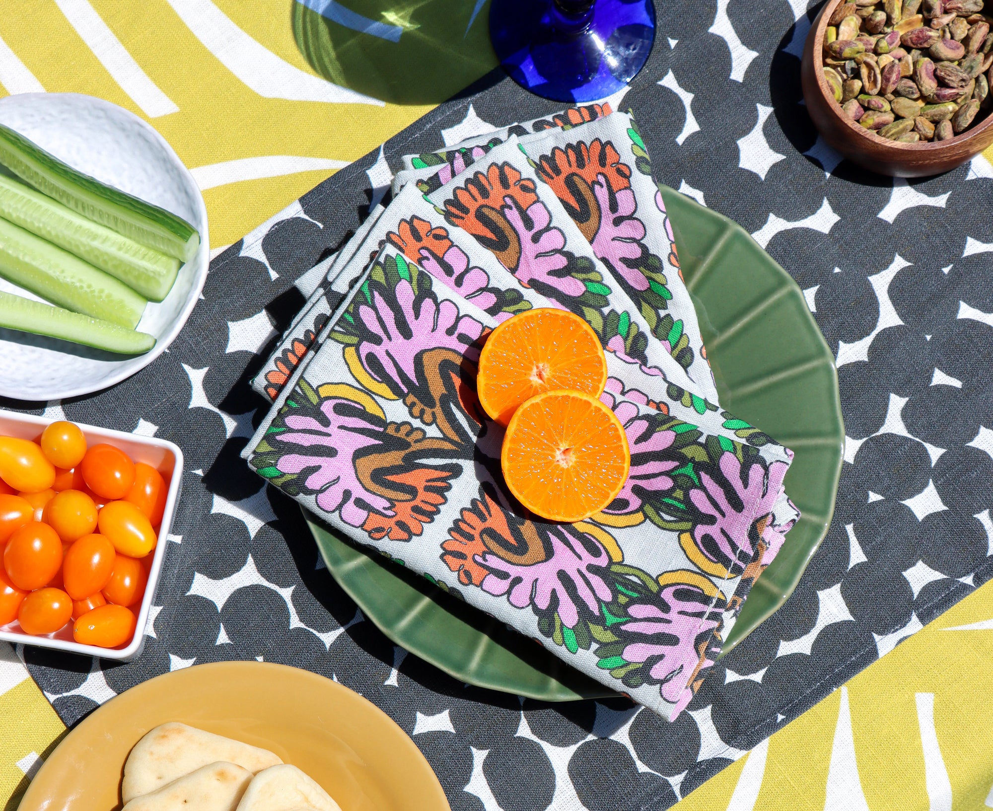 Colorful layered table linens designed by artist Leila Simon Hayes. Floral napkins with orange slices on a patterned black and white dots placemat, on a yellow striped tablecloth with snacks around.