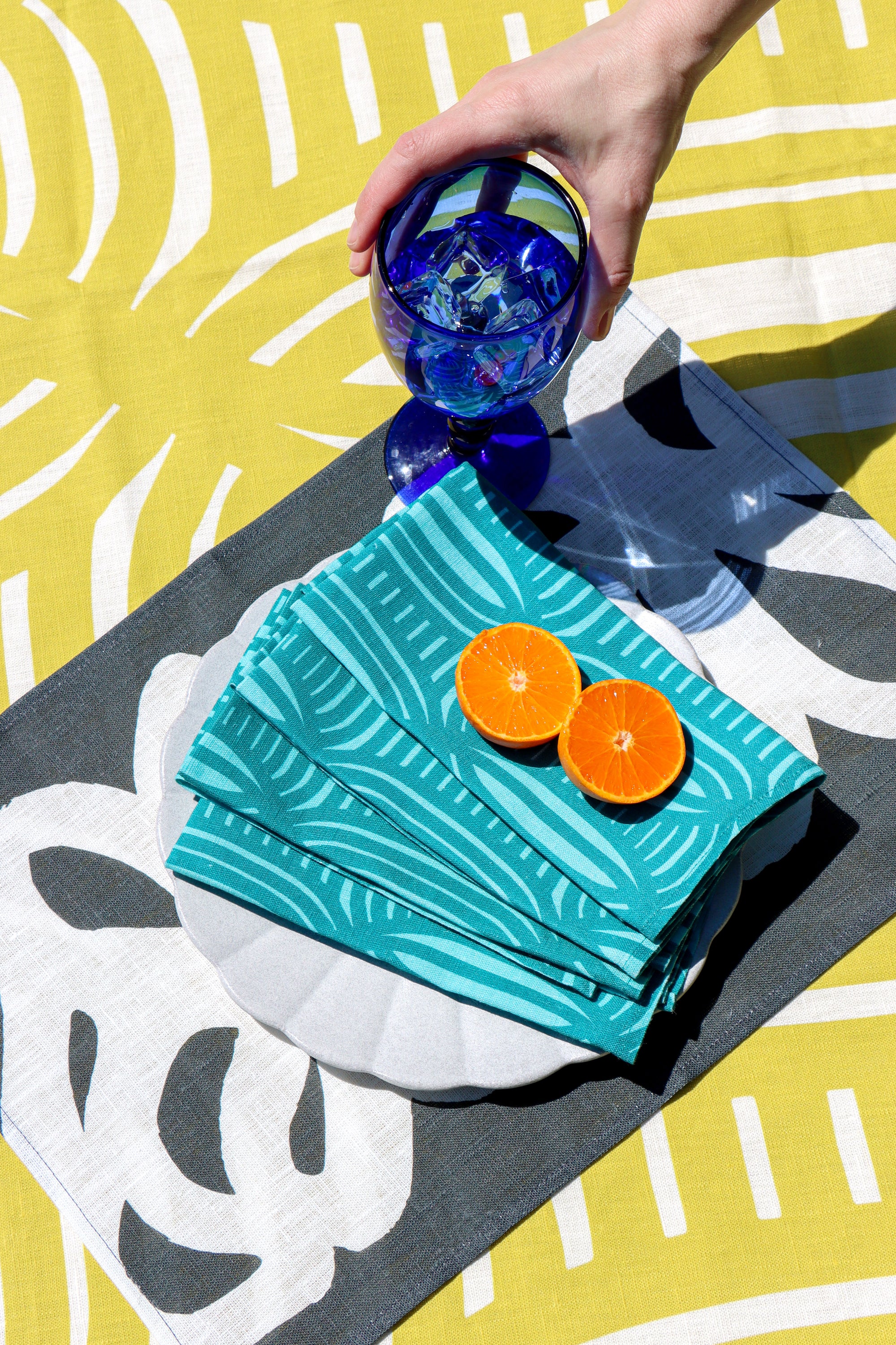 A lifestyle shot of a place setting featuring a stack of teal and white patterned paper napkins on a white plate, garnished with two orange slices, set on a gray placemat Over a yellow and white patterned tablecloth. Designed by artist Leila Simon Hayes.