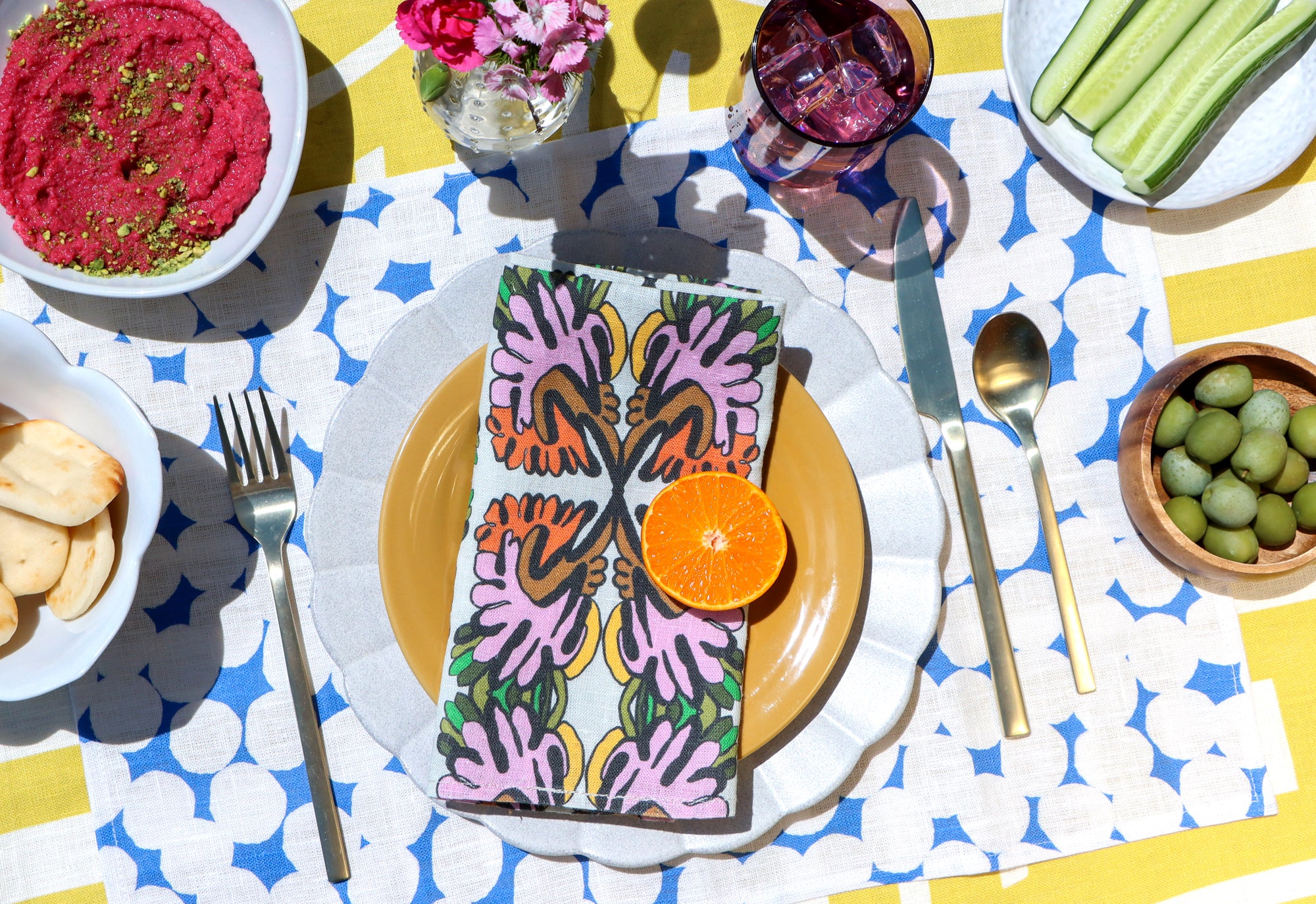 Colorful layered table linens designed by artist Leila Simon Hayes. Floral napkins on a patterned blue and white dots placemat, on a yellow striped tablecloth with snacks around.