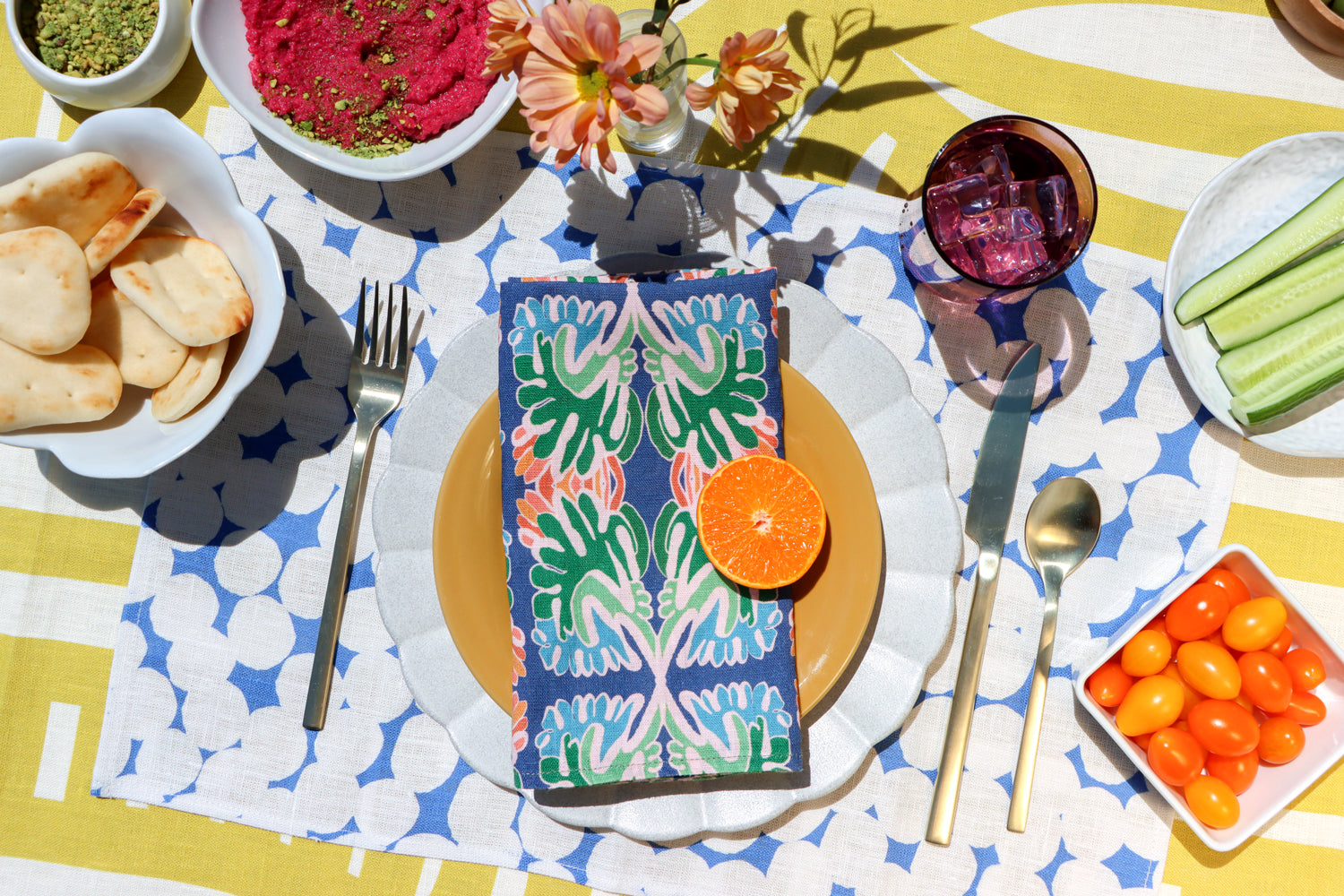 Colorful layered table linens designed by artist Leila Simon Hayes. Blue floral napkins with orange slices on a patterned blue and white dots placemat, on an orange striped tablecloth with snacks around.