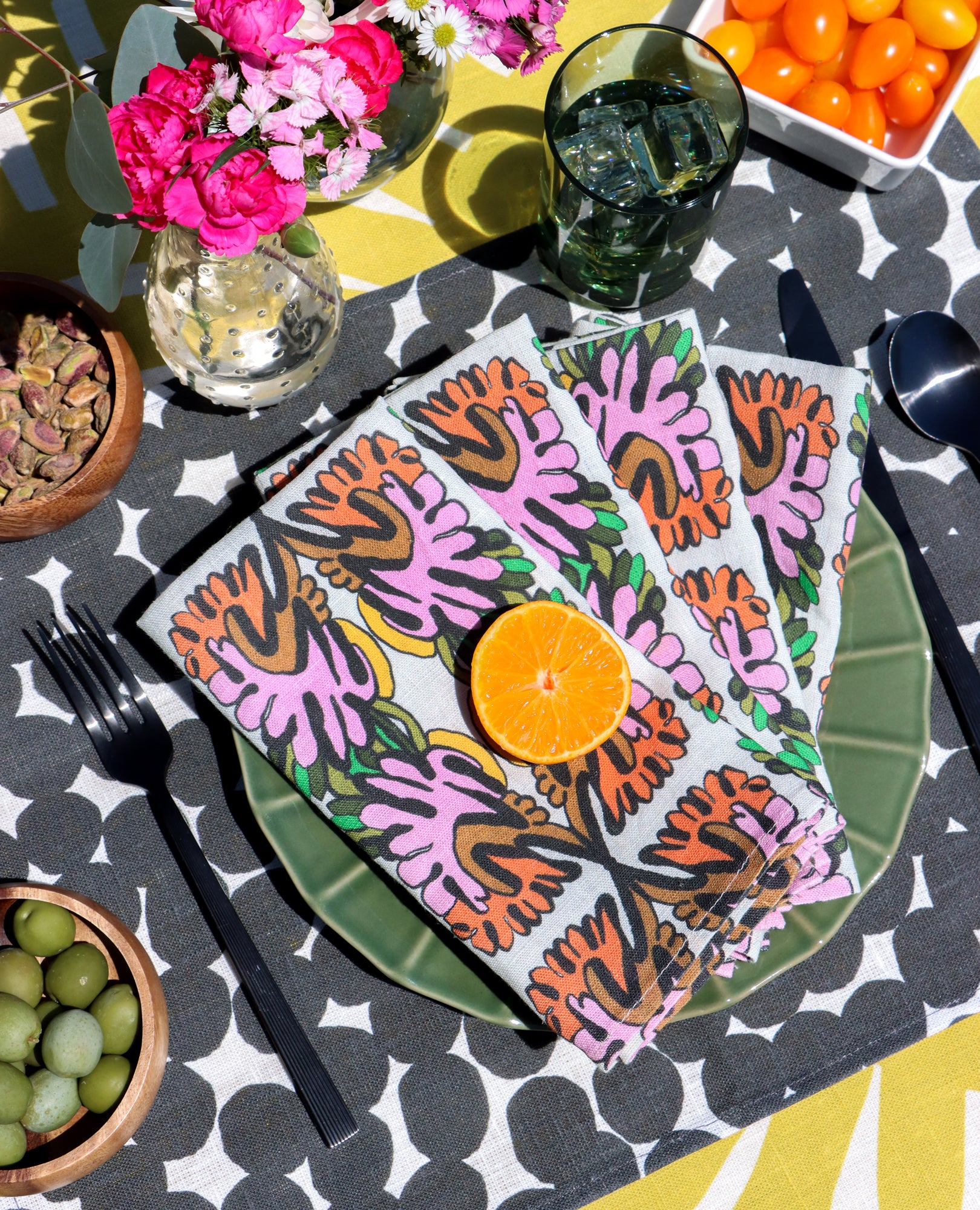 Colorful layered table linens designed by artist Leila Simon Hayes. Floral napkins with orange slices on a patterned black and white dots placemat, on a yellow striped tablecloth with snacks around.
