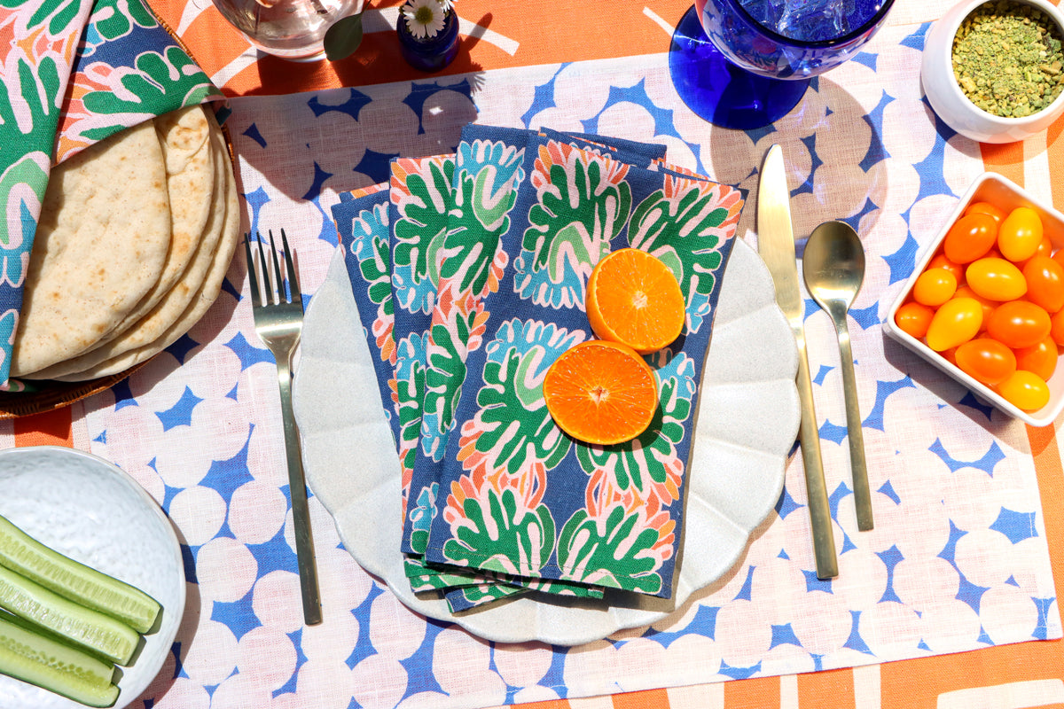Colorful layered table linens designed by artist Leila Simon Hayes. Blue floral napkins with orange slices on a patterned blue and white dots placemat, on an orange striped tablecloth with snacks around.