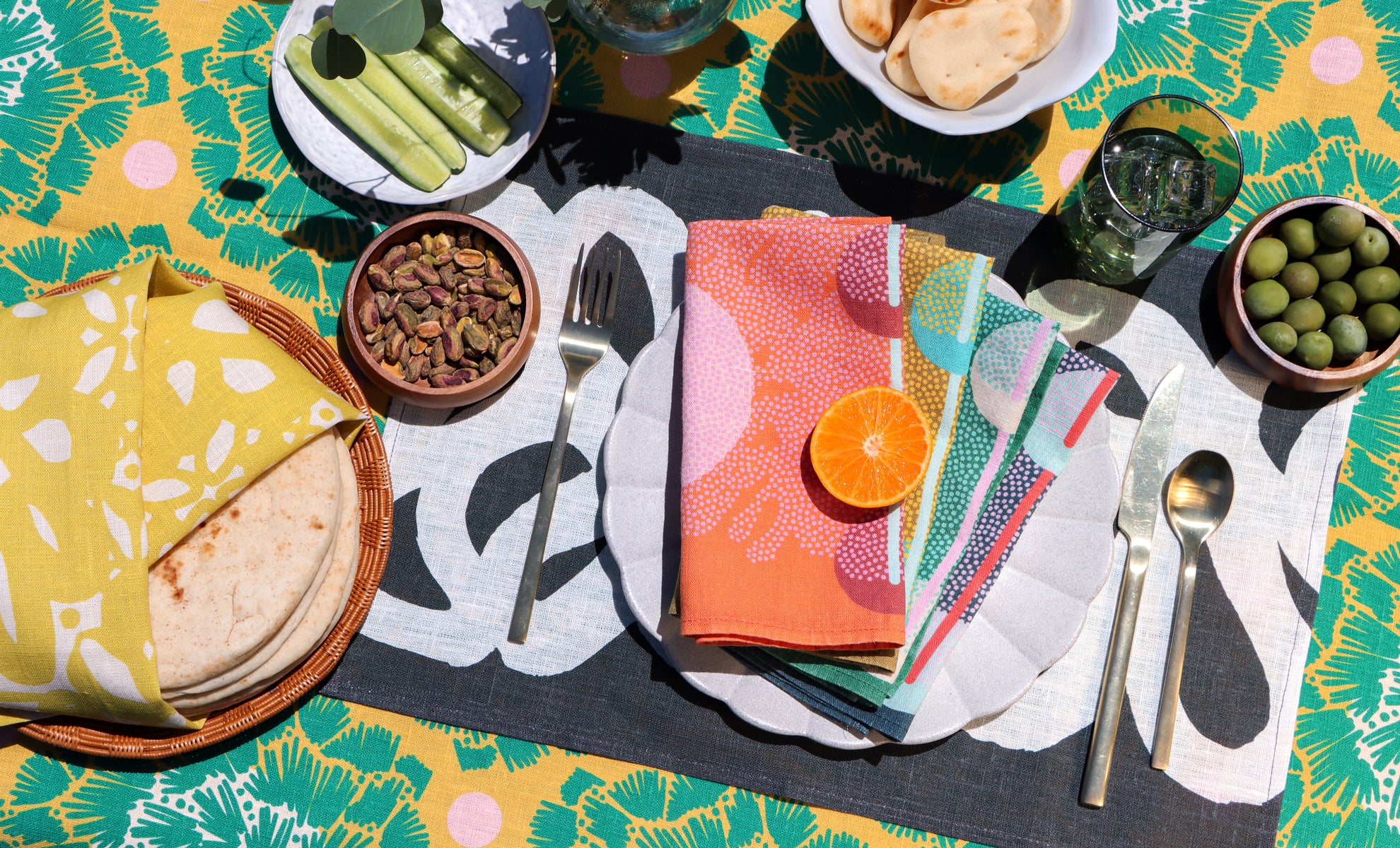 A styled outdoor dining table setting with a vibrant green and yellow palm-patterned tablecloth, a black placemat, patterned napkins, a stack of tortillas, pickled vegetables, olives, and a slice of orange. Designed by artist Leila Simon Hayes.