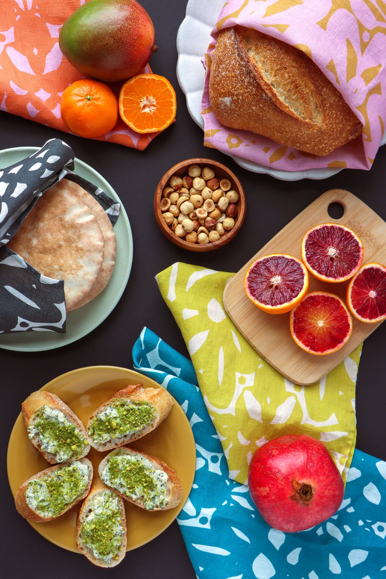 Assorted fruits, bread, and nuts on a dark surface with colorful tablecloths. Designed by artist Leila Simon Hayes.